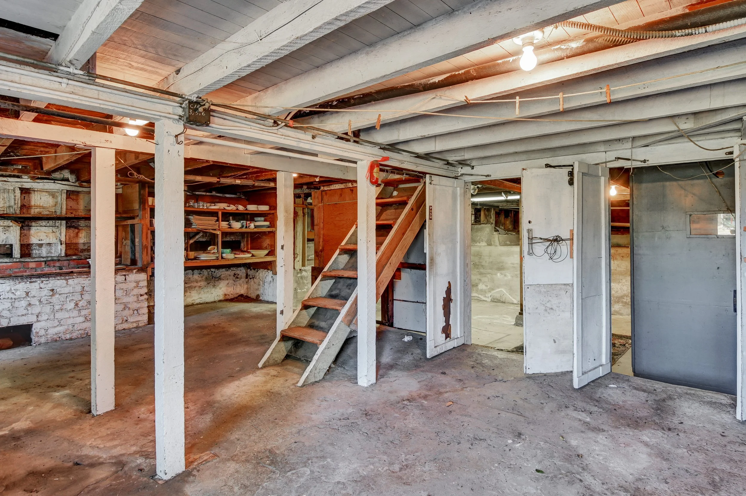 Interior of a basement with exposed beams, brick walls, shelves with dishes, and a set of wooden stairs.