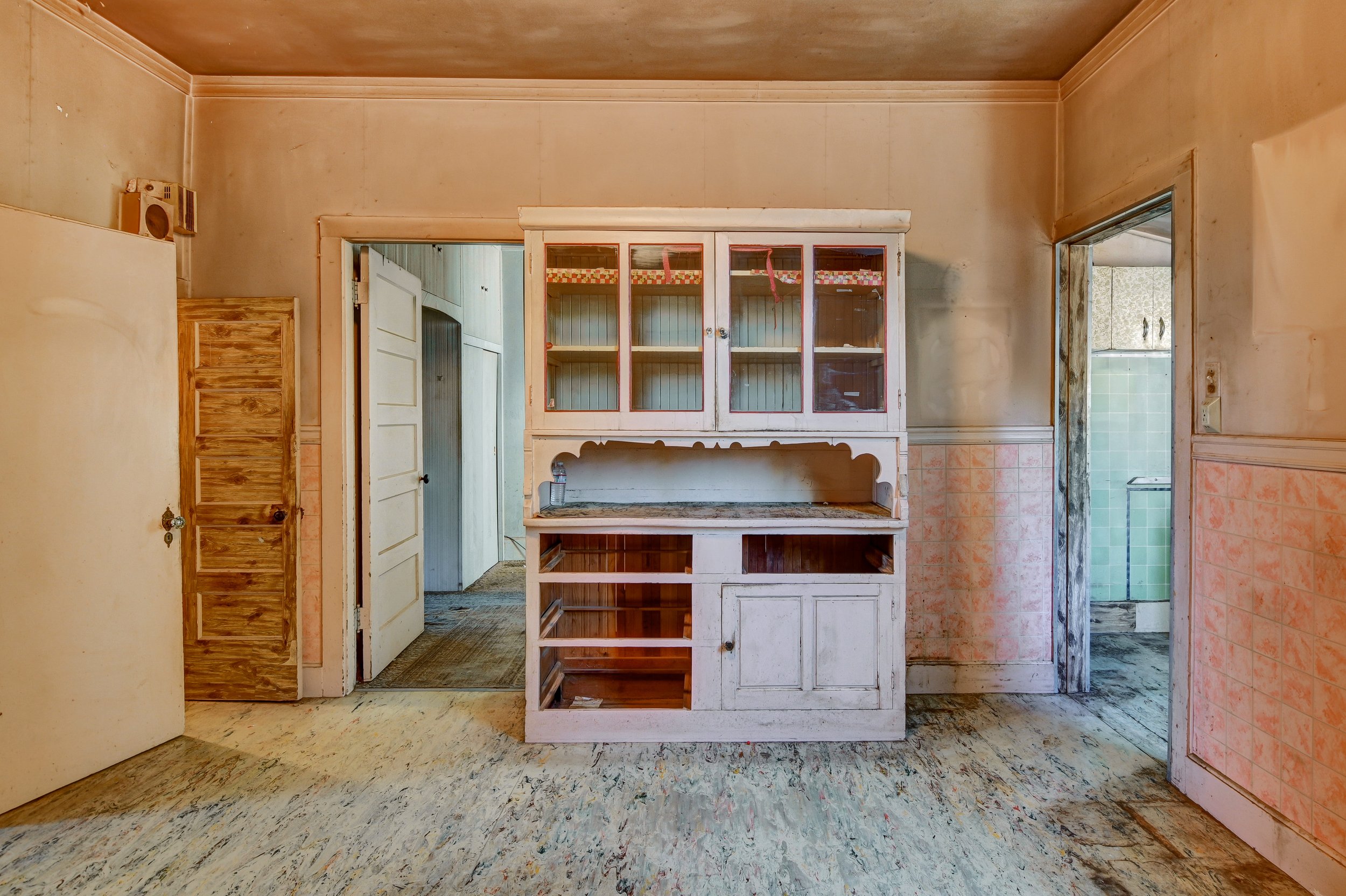 Empty kitchen with an old white cabinet with glass doors, open shelves, and a granite countertop. Walls are faded pink and beige with peeling paint. The floor shows worn, stained wooden planks, and there are open doorways leading to other rooms, incl