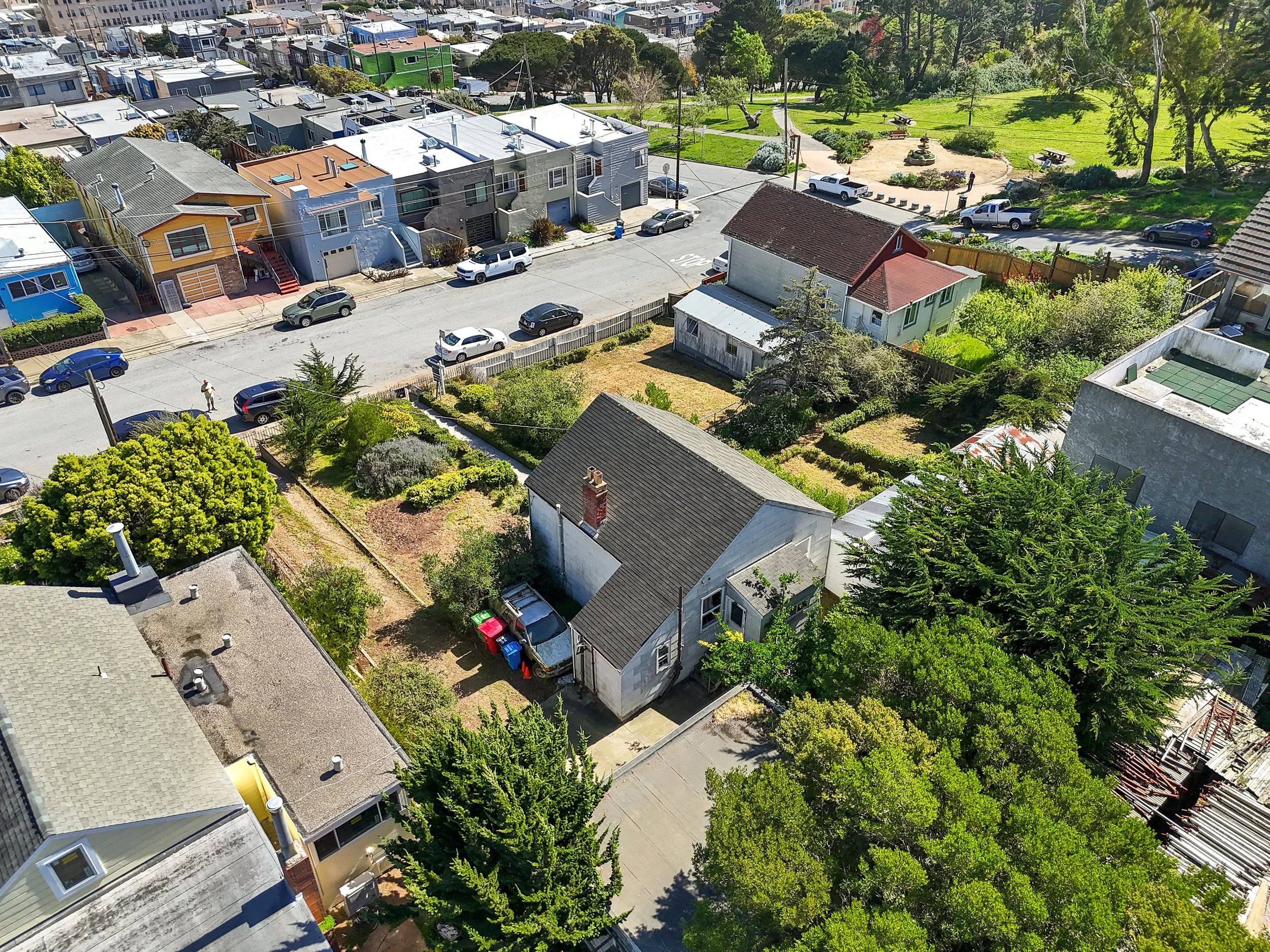 An aerial view of a residential neighborhood featuring houses, a green park with walking paths and benches, and a street with cars parked along the curb.