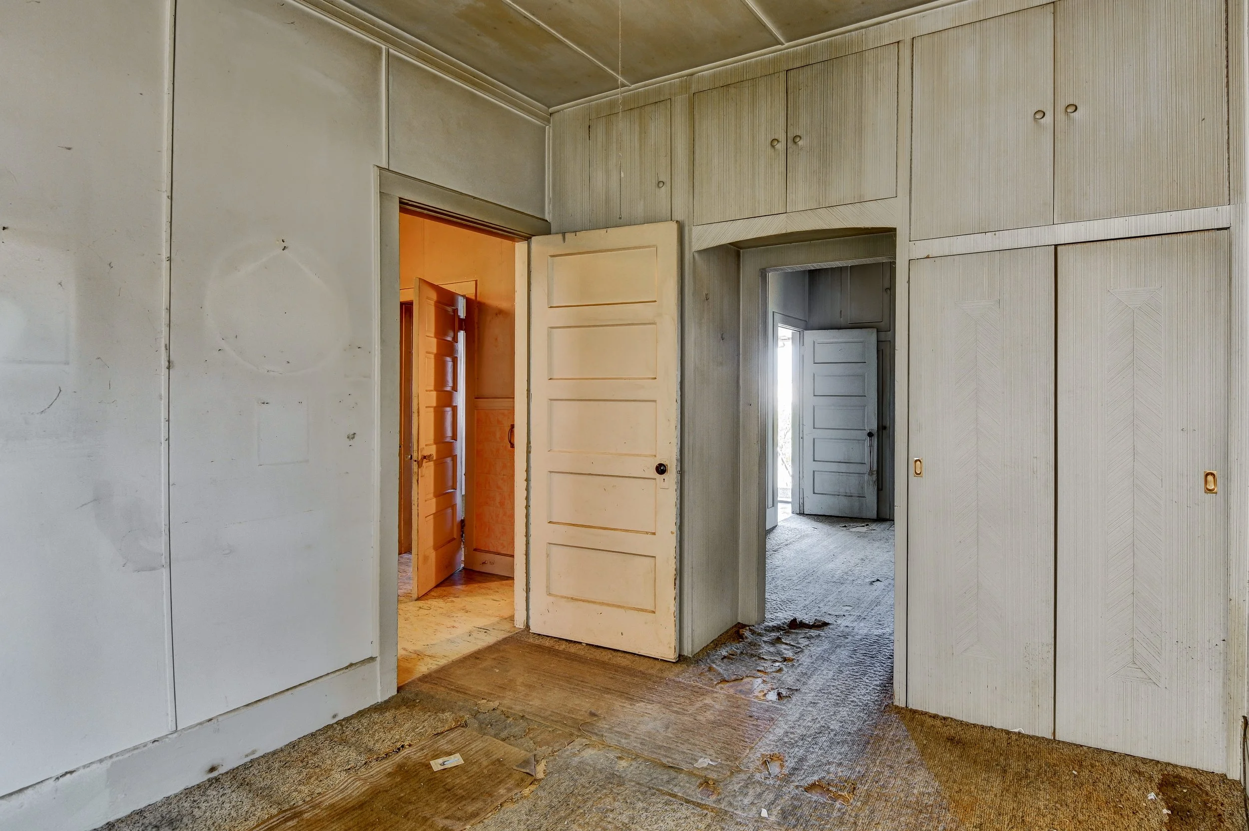 Interior of an old, abandoned house with worn carpet and peeling floors, featuring multiple open doors leading to other rooms, with faded wood paneling and a cluttered, dusty appearance.