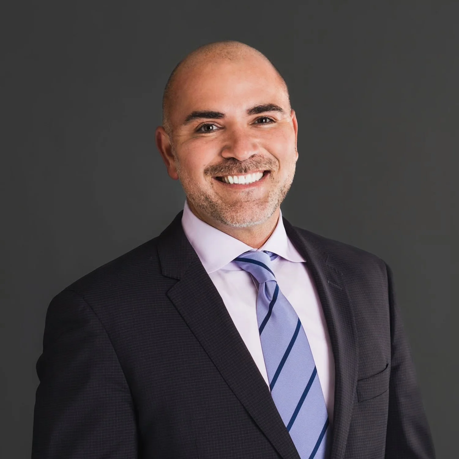 A professional headshot of a smiling man with a shaved head, wearing a dark suit, light purple shirt, and striped blue tie, against a dark gray background.