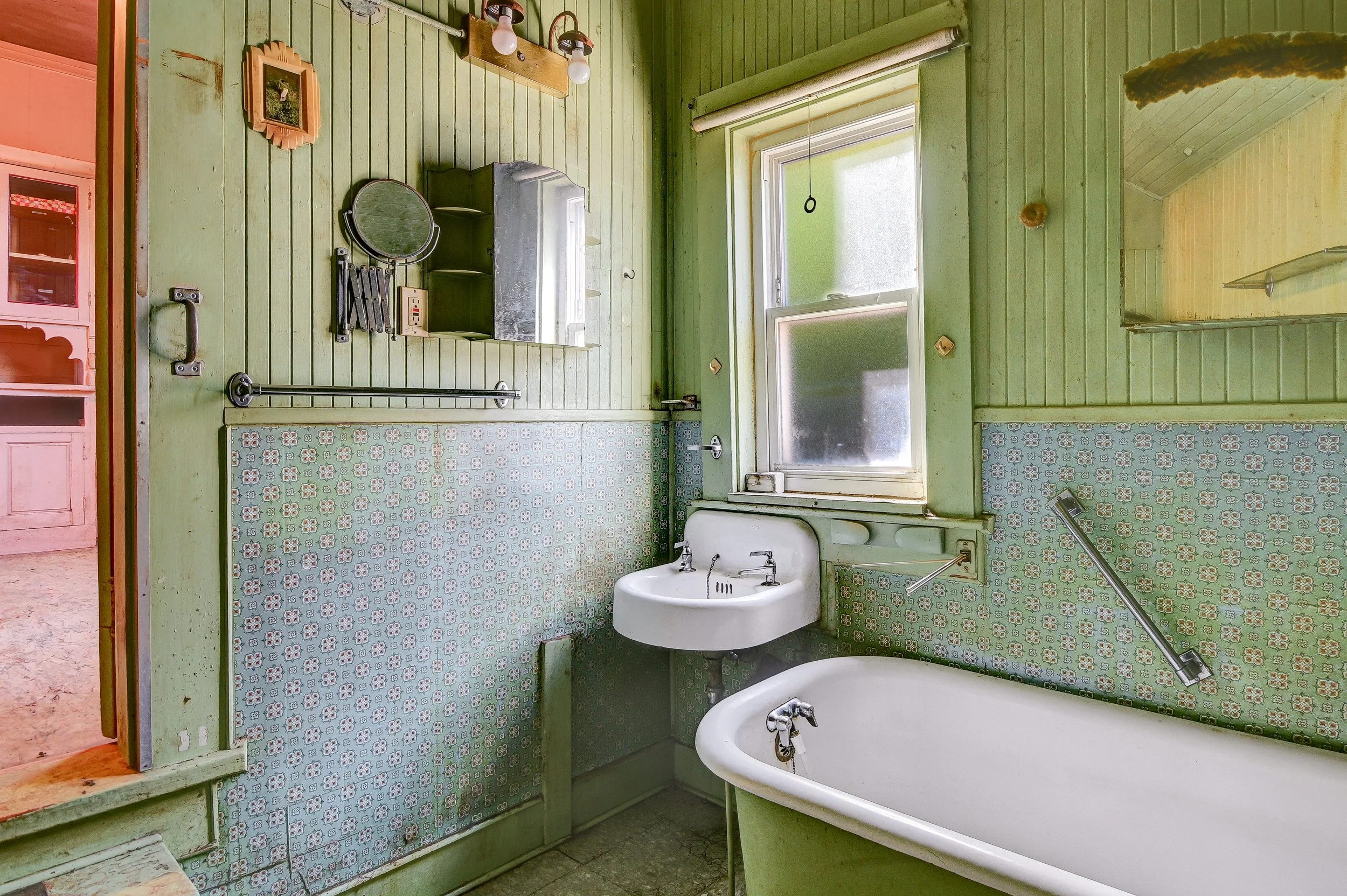 Old-fashioned bathroom with green vertical wood-paneled walls, patterned tile halfway up, a small white sink, a bathtub, and a window with frosted glass.