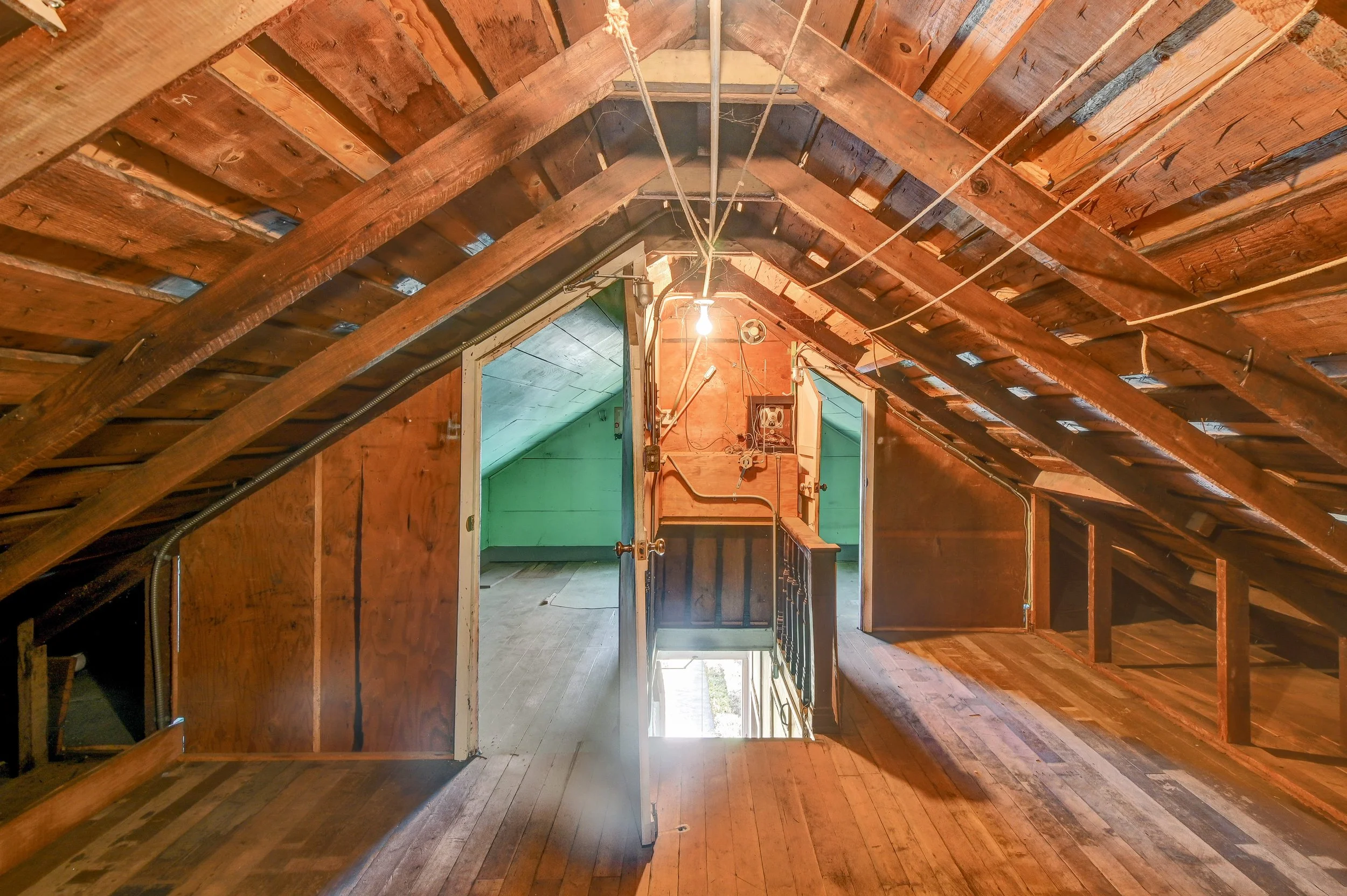 An interior view of a rustic attic with wooden floors and slanted ceilings, a staircase leading down, and a green wall in the background.