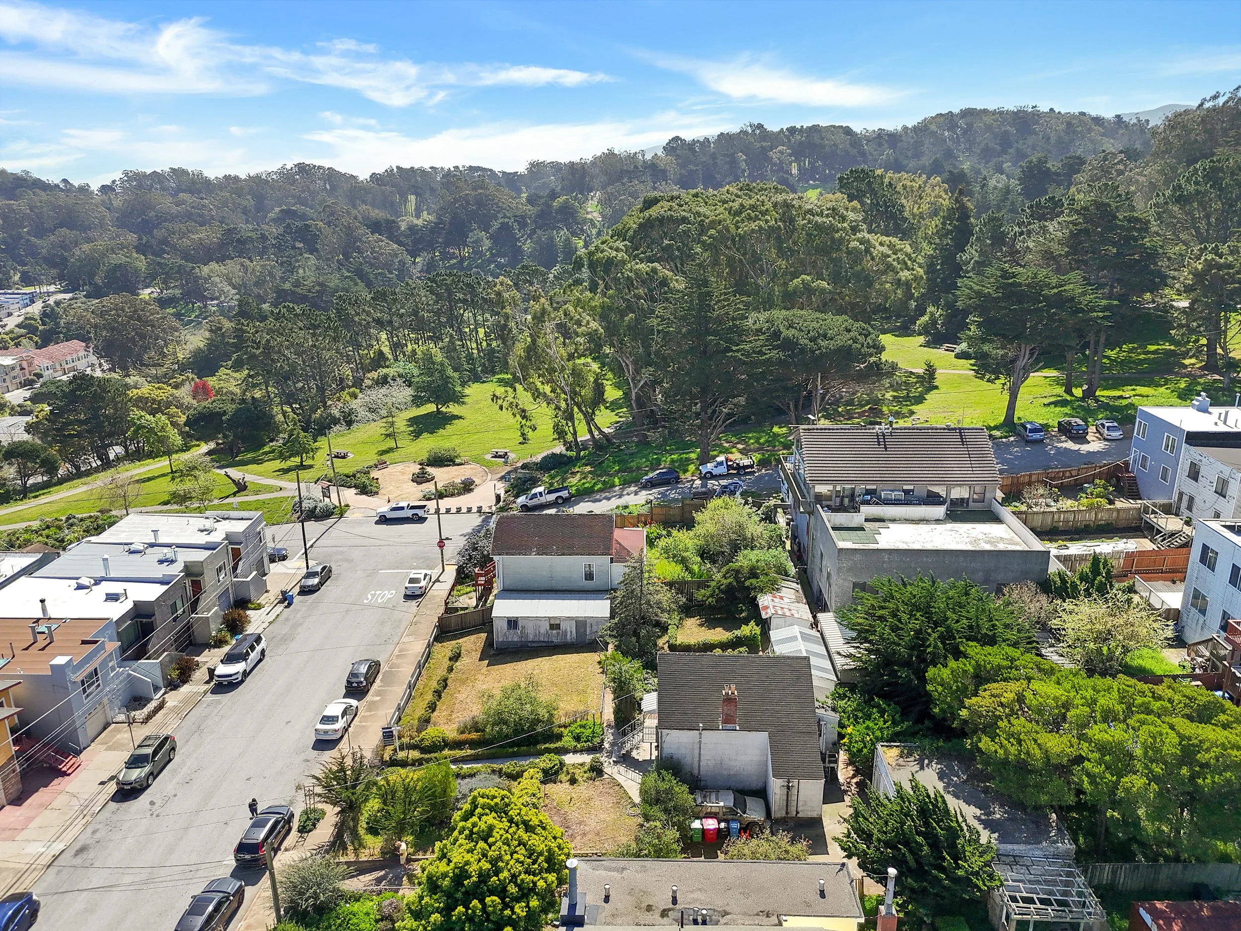 An aerial view of a neighborhood with houses, trees, a park, and a parking lot under a clear blue sky.