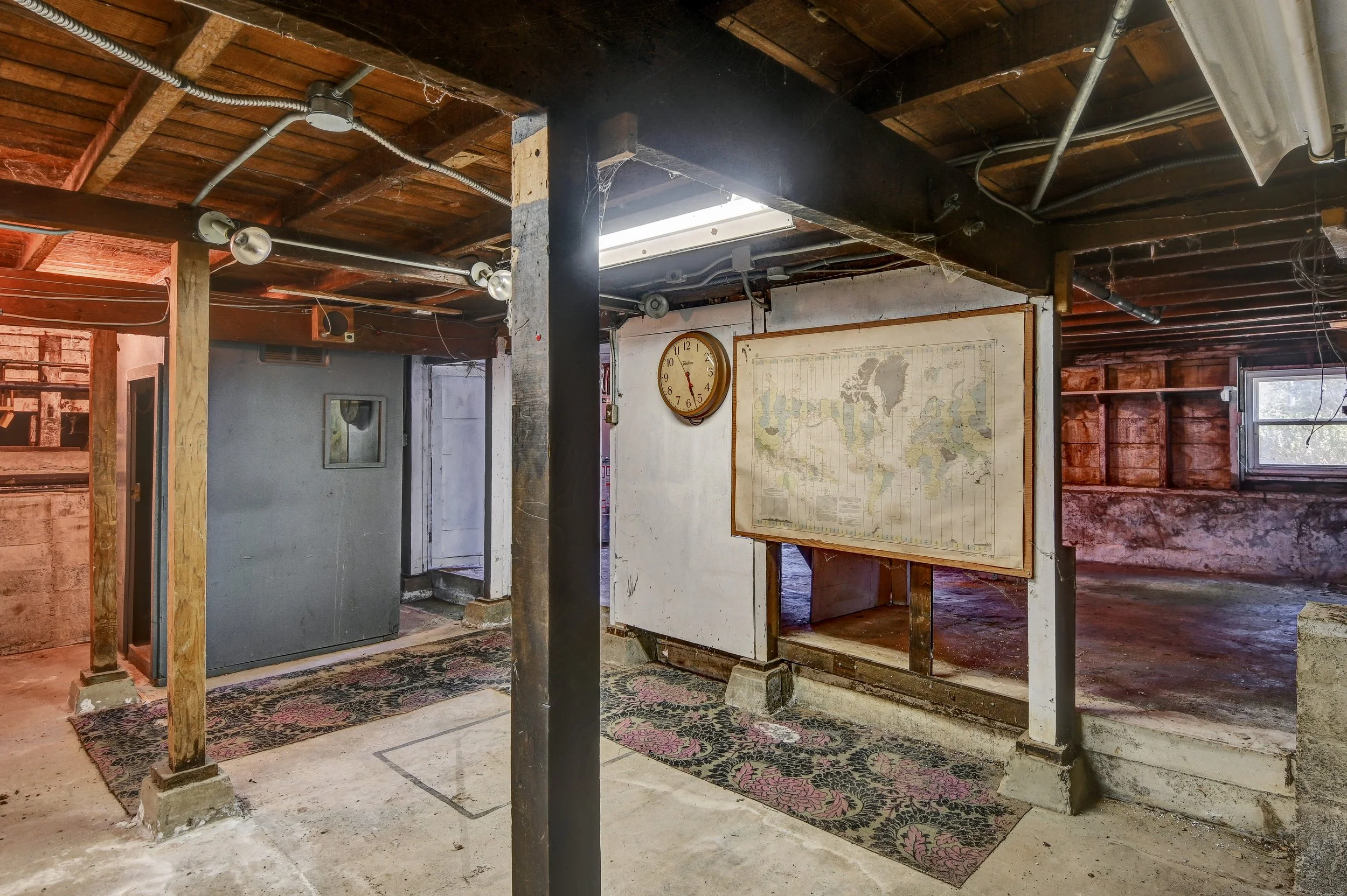 Interior of a basement or garage with exposed wooden ceiling beams, a white wall with a world map, an old wall clock, and a small window.