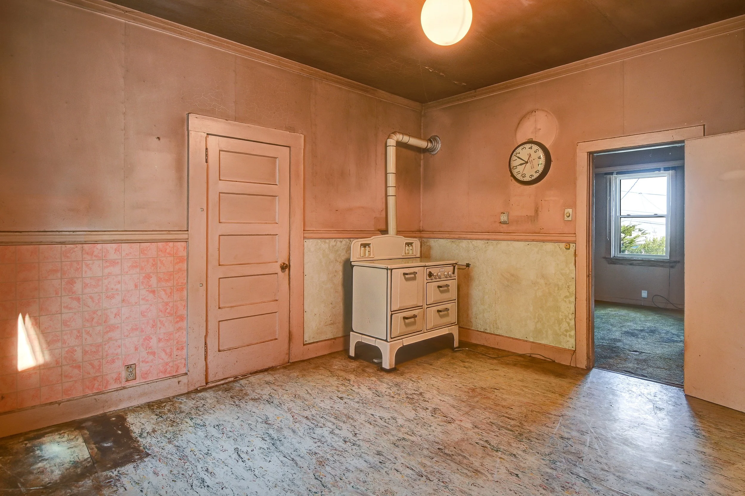 Empty kitchen with worn pink walls, a vintage stove, a window showing outside greenery, and a wall clock.