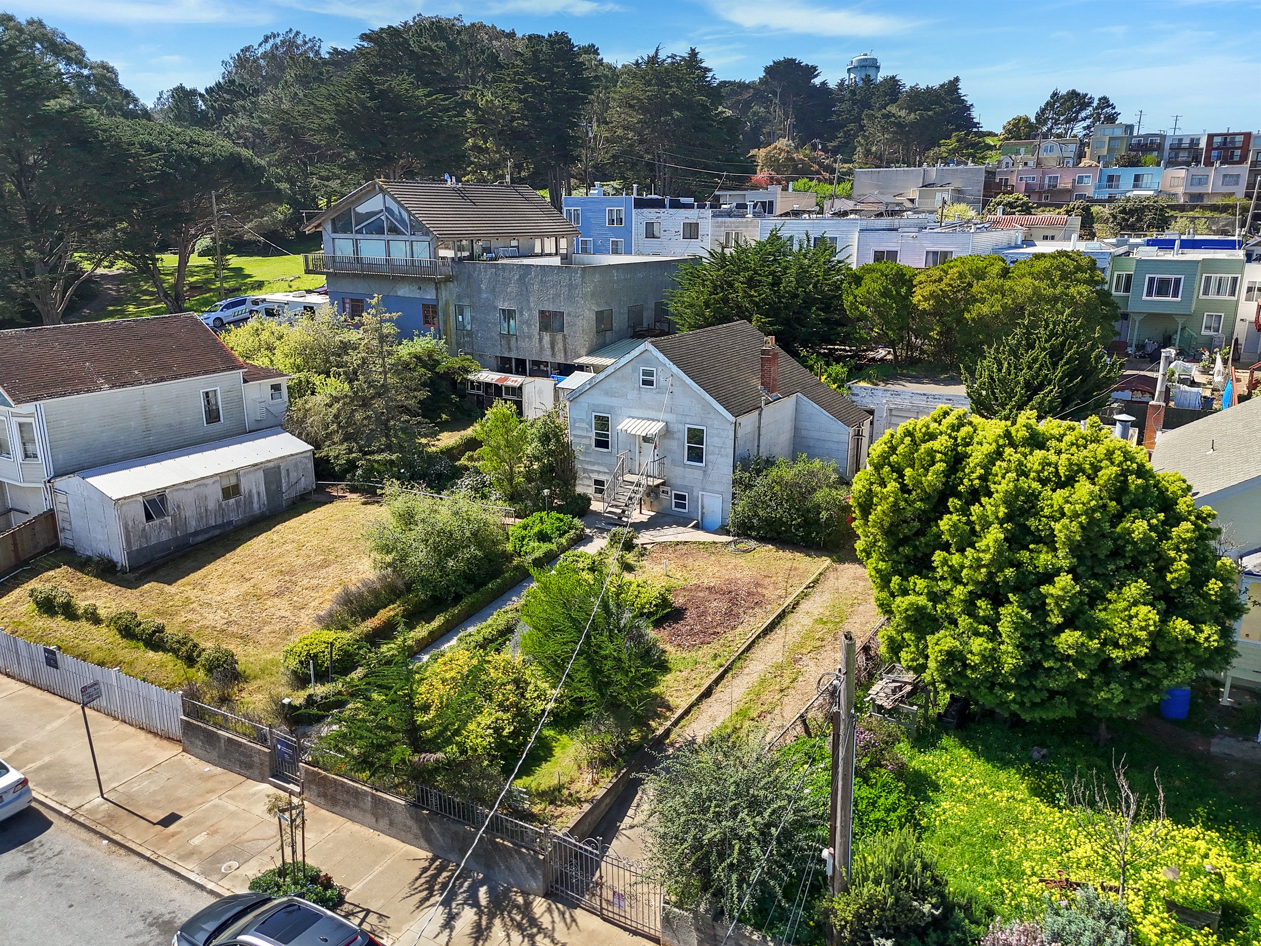 Aerial view of a residential neighborhood with a small backyard, large tree, and houses on a hillside, with cars parked along the street.