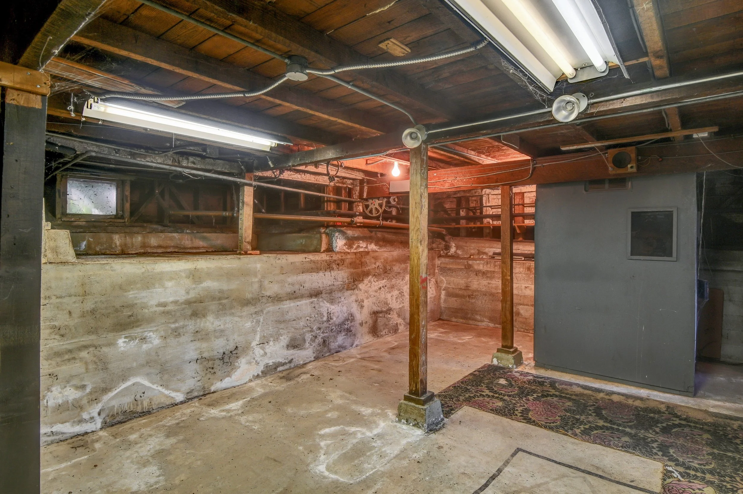 Empty basement with exposed pipes, wooden support beams, a small window, and a gray utility box on the wall. The floor is concrete with some discoloration and a patterned rug.