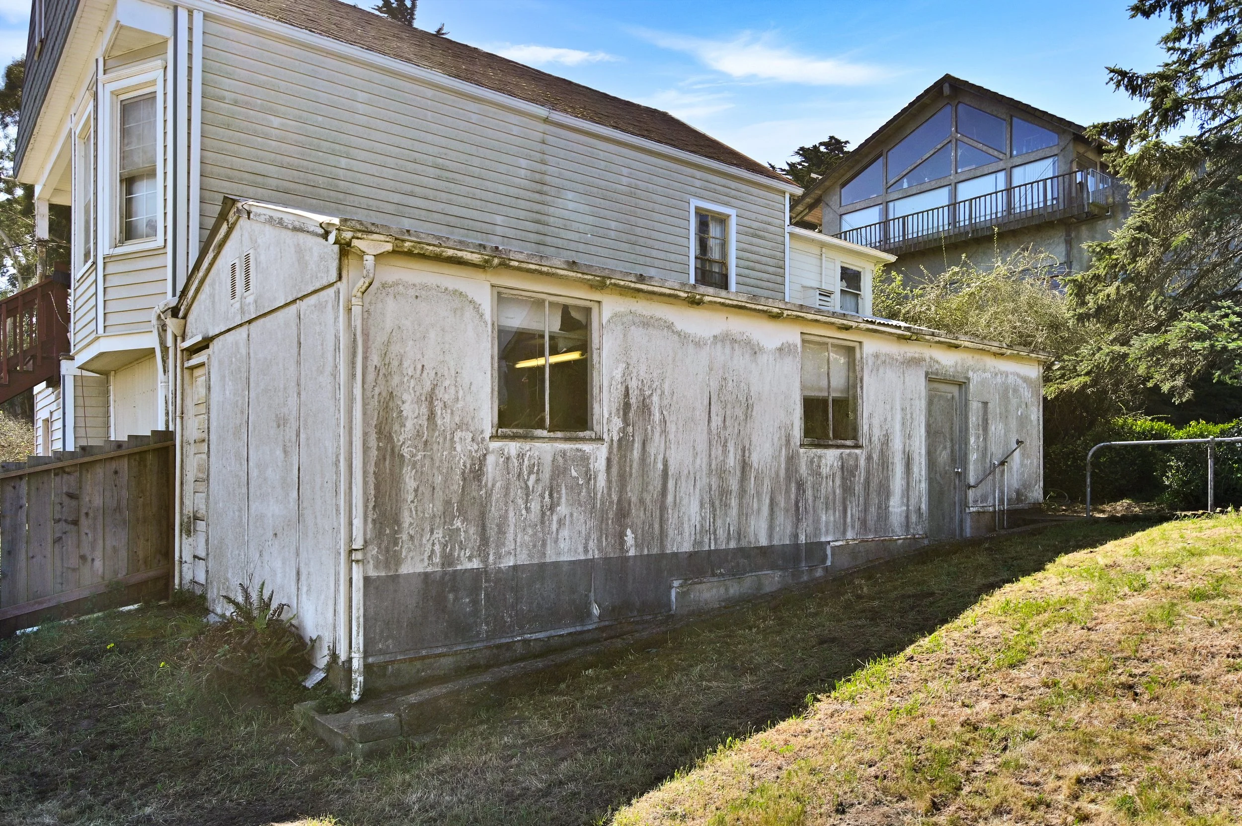 Backyard of a house with dirty, weathered walls, two windows, a security door, and a side ramp, with an elevated house with large windows and a balcony in the background.