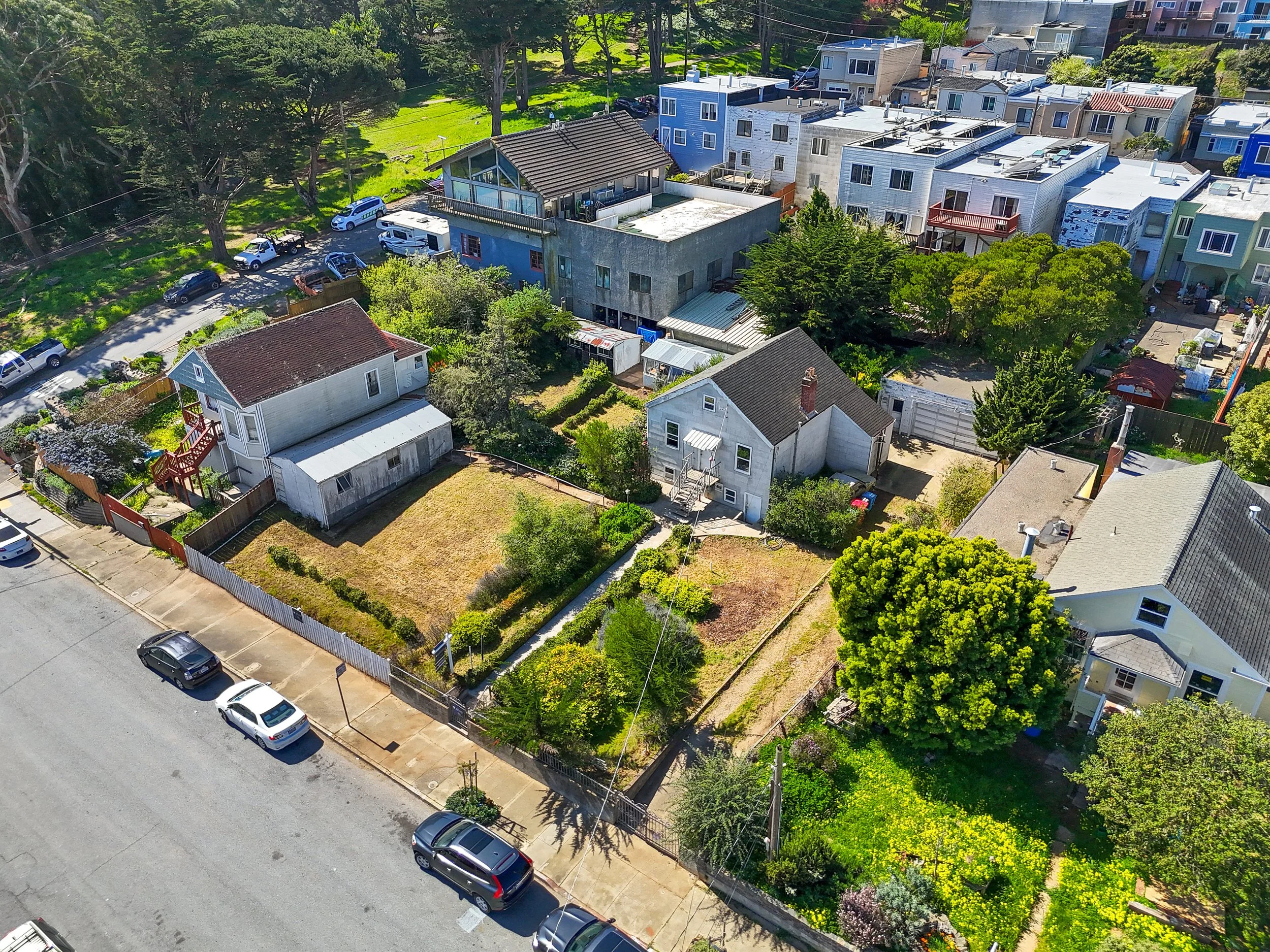 An aerial view of a neighborhood with several houses, some with yards, trees, and parked cars. There are multi-story buildings in the background and a street with cars parked along the curb in the foreground.