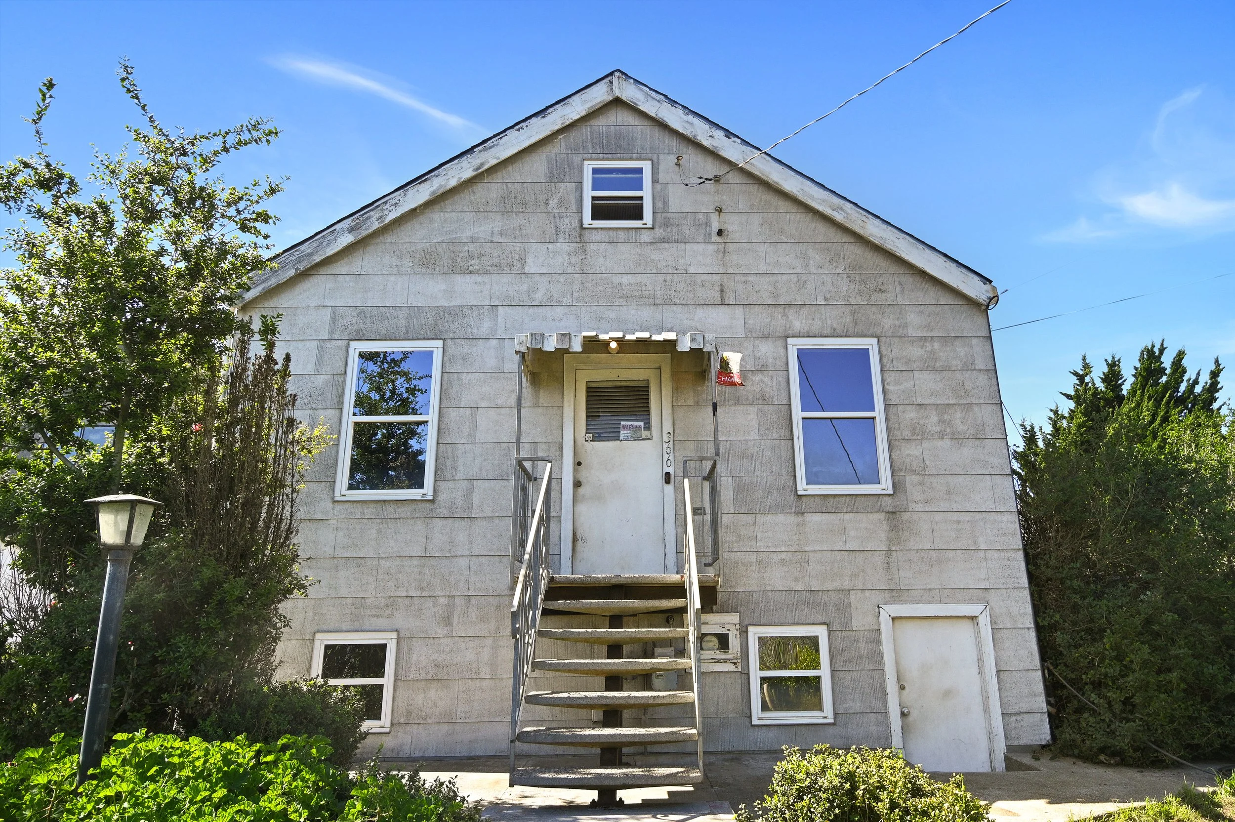 A two-story house with gray siding, six windows, a front door with a small covered porch, and a staircase leading up to the door. There are bushes and a streetlamp in front, with a clear blue sky above.