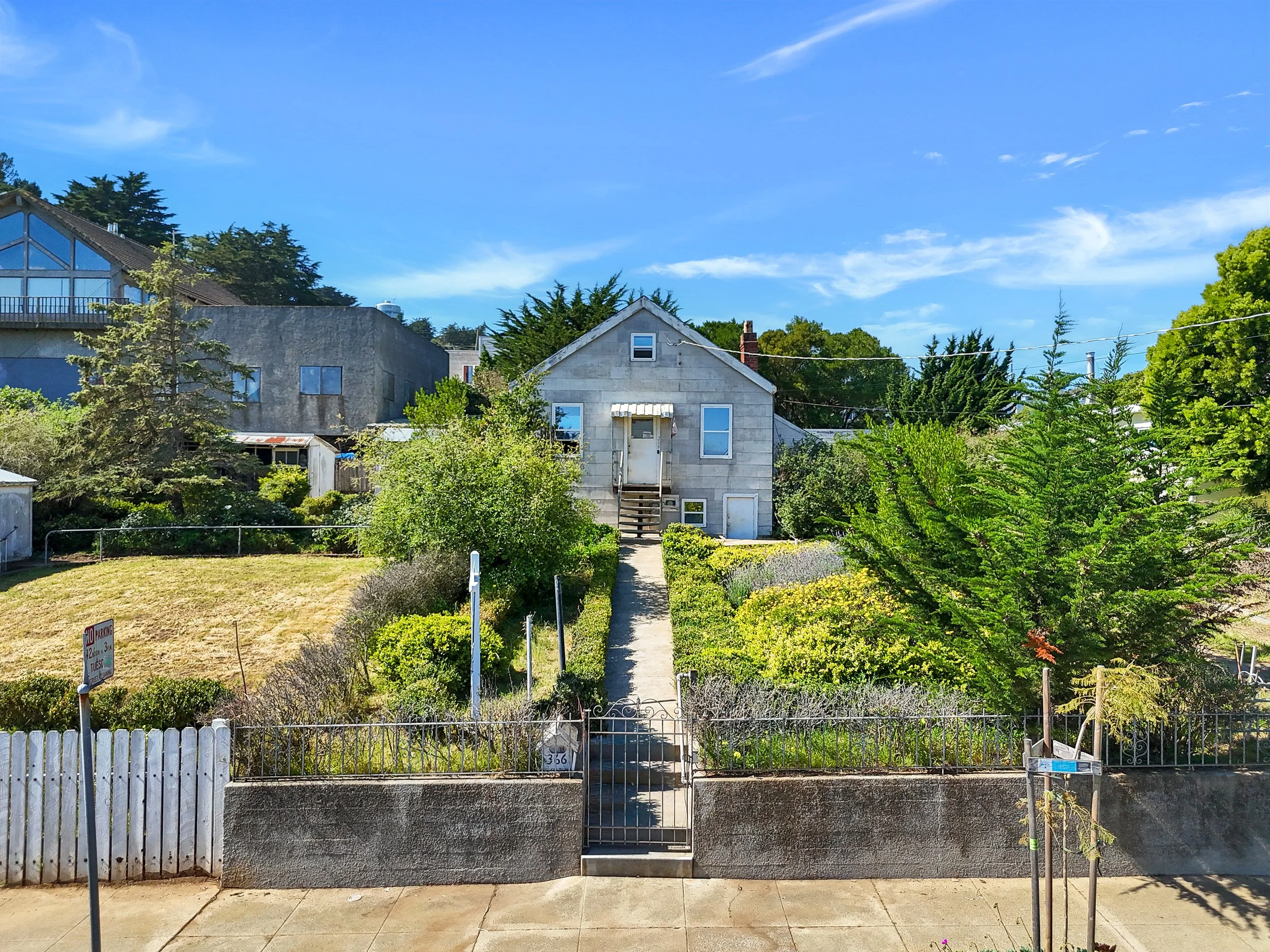 Residential house with a front garden, trees, and a sidewalk under a blue sky with clouds.