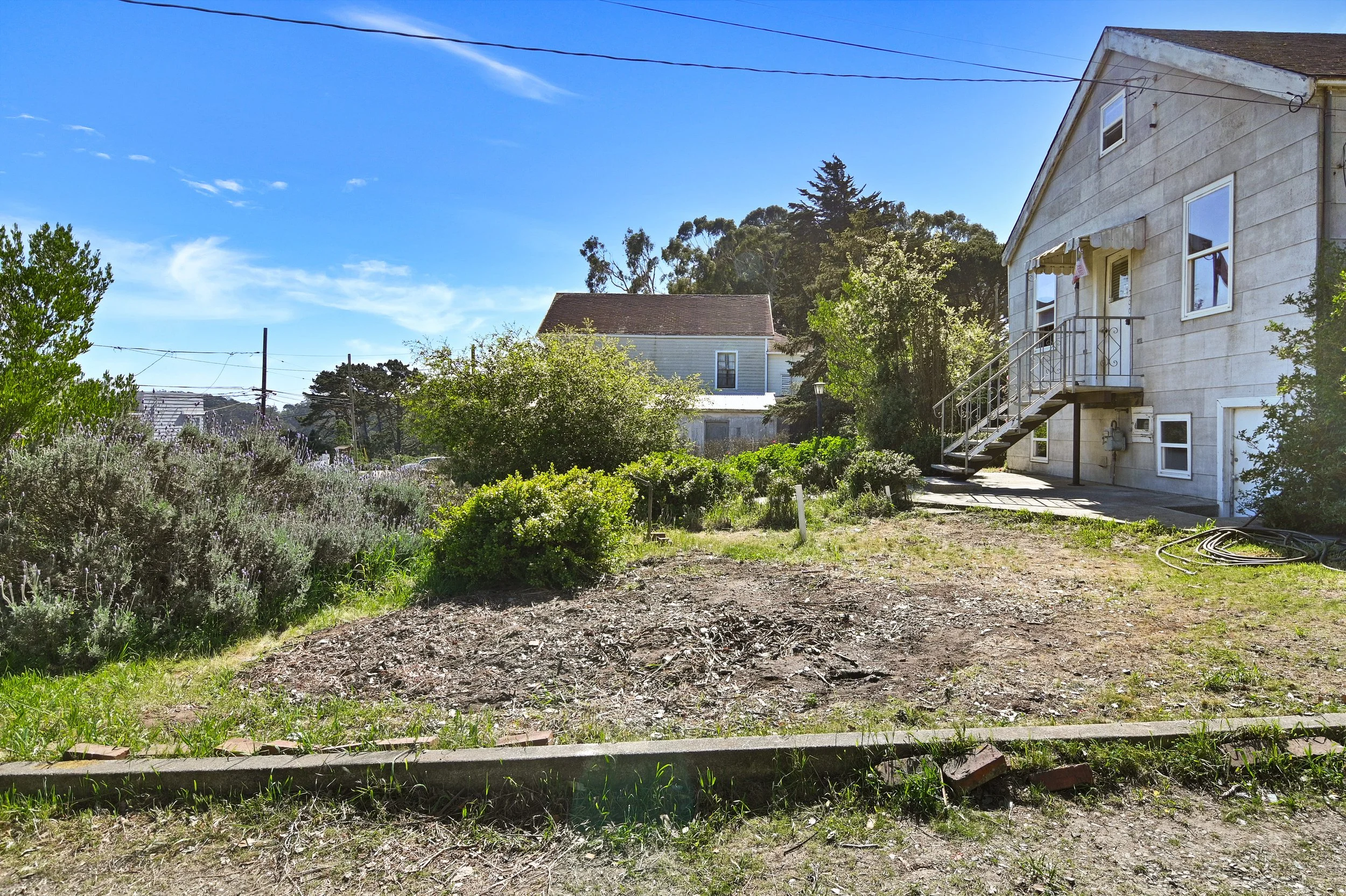 A residential backyard on a sunny day with a patch of dirt, green bushes, and an outdoor staircase leading to the second floor of a house. The house has beige siding, a window with curtains, and nearby garden hoses.