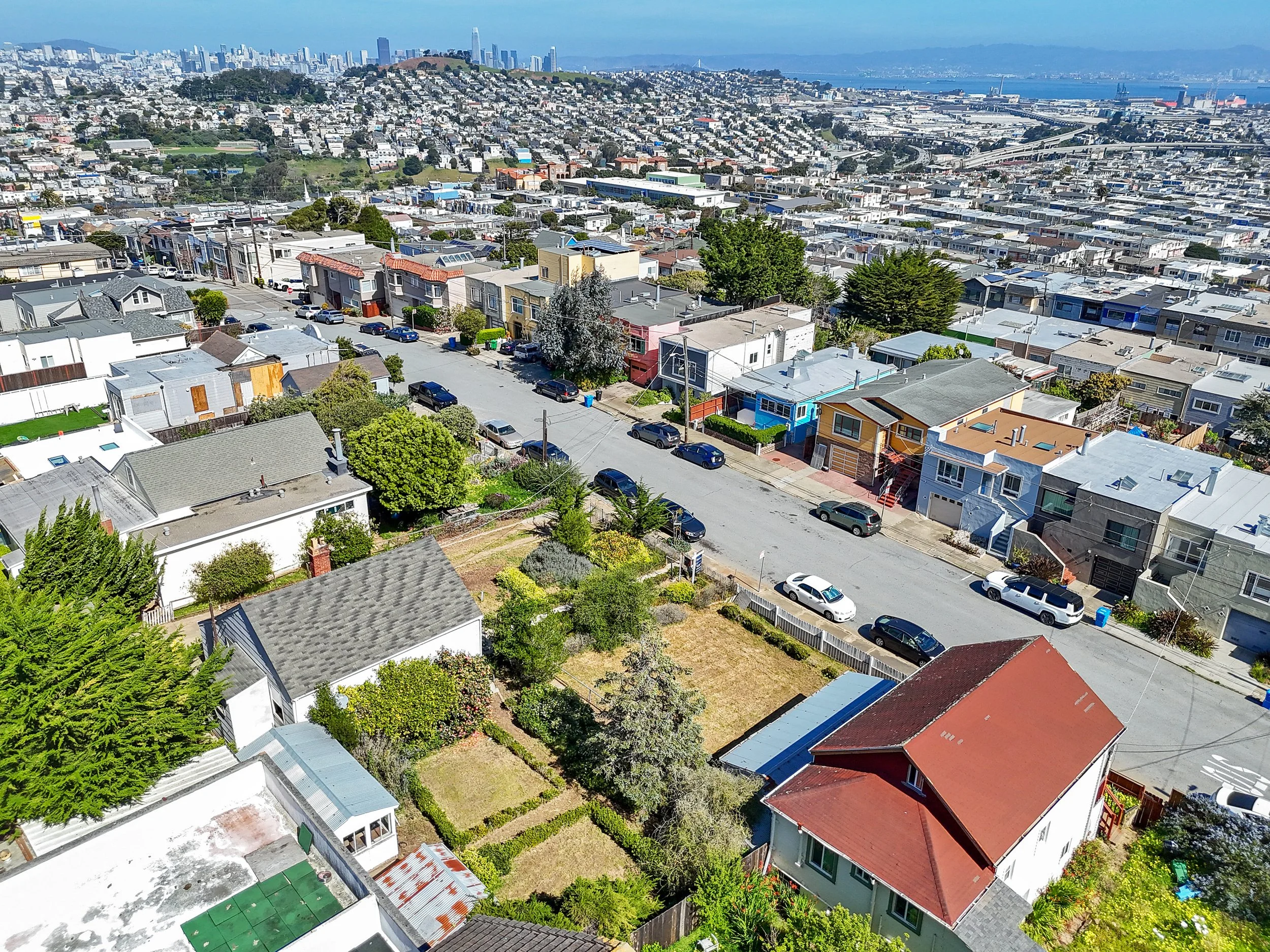 An aerial view of a residential neighborhood with houses, trees, and parked cars, with a cityscape and bay in the background.