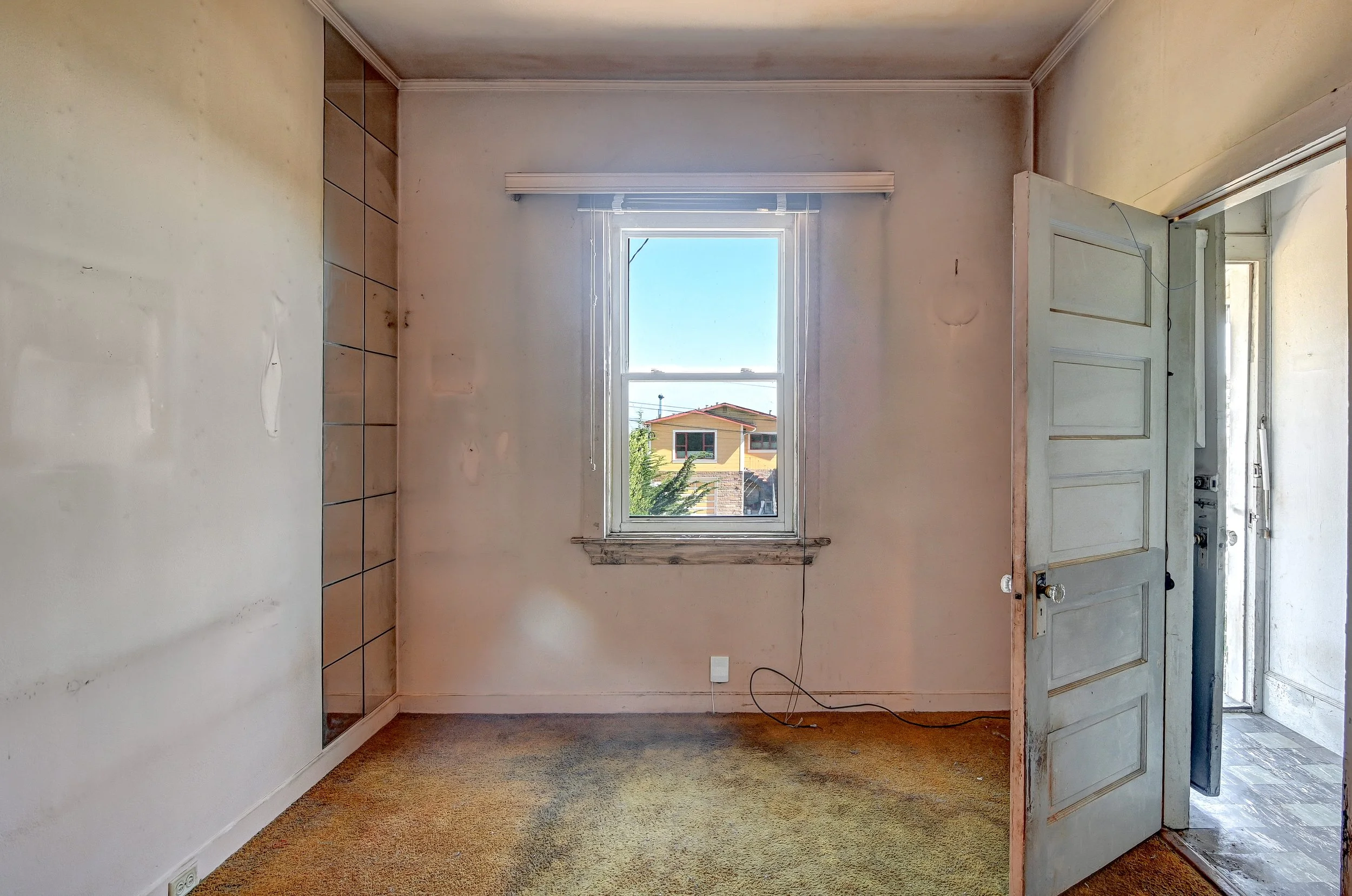 Empty room with a window revealing a yellow house outside, worn carpeting, and an open door leading to a hallway.