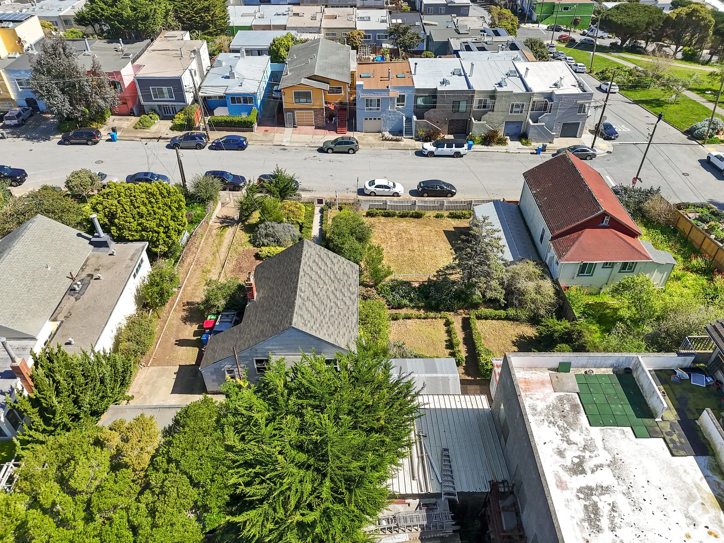 Aerial view of residential neighborhood with houses, parked cars along the street, and backyard gardens, with a row of colorful houses and lush green trees.
