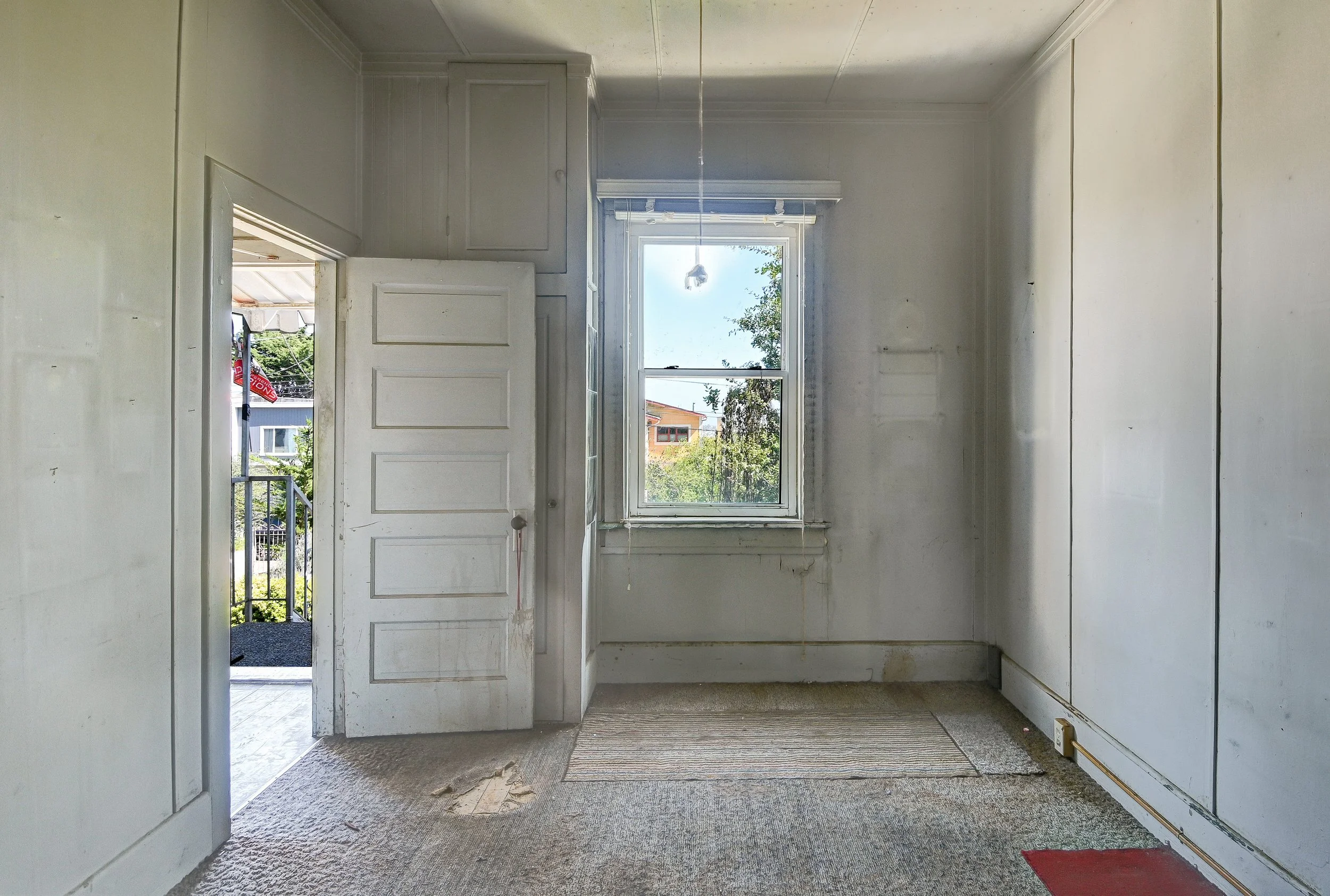 Empty room with a window showing trees and buildings outside, partially open door leading to an outdoor area, beige carpeted floor with stains, white walls, and an outlet near the floor.