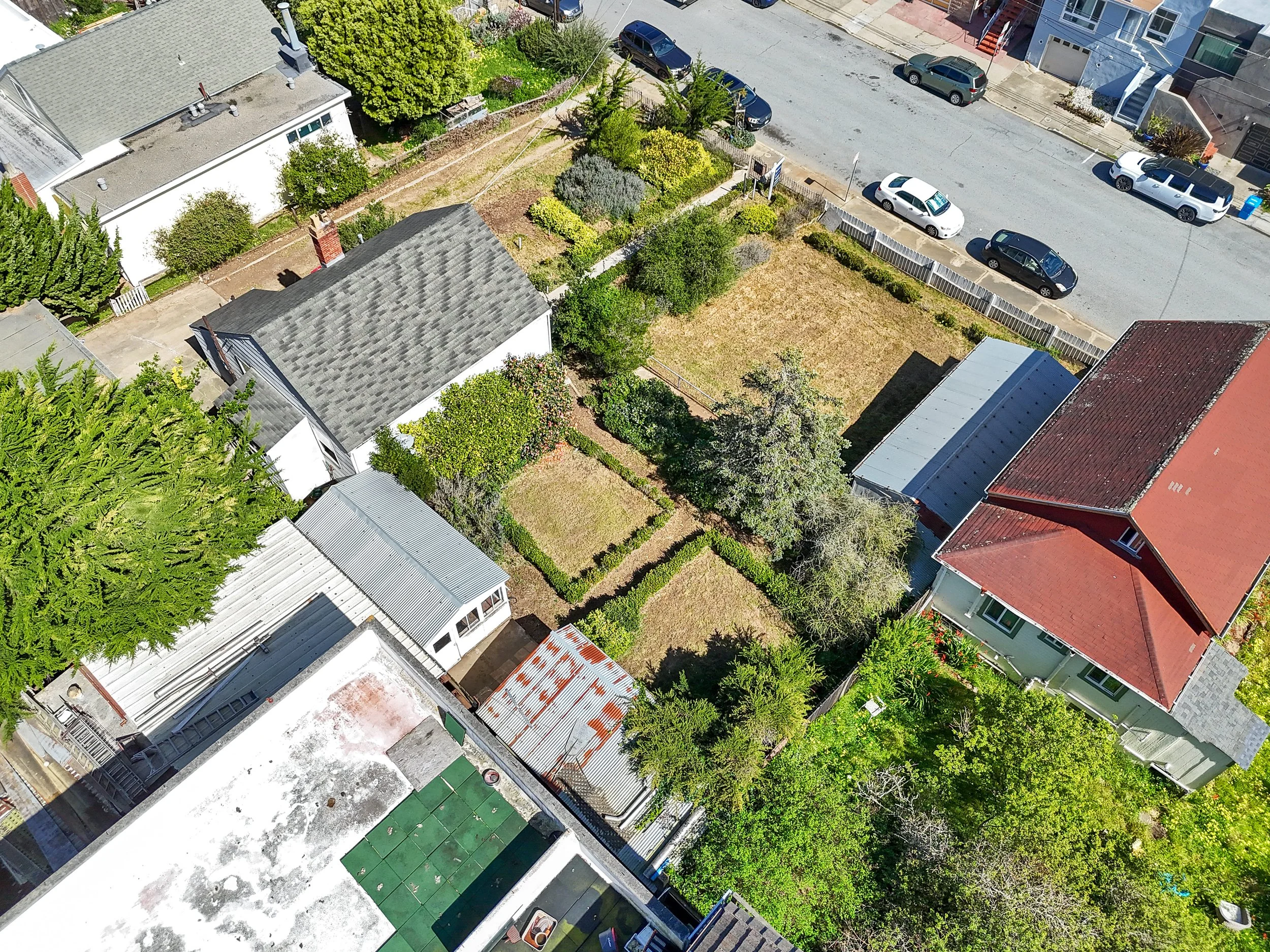 Aerial view of a residential neighborhood showing houses, a street with parked cars, and backyard gardens with trees and bushes.