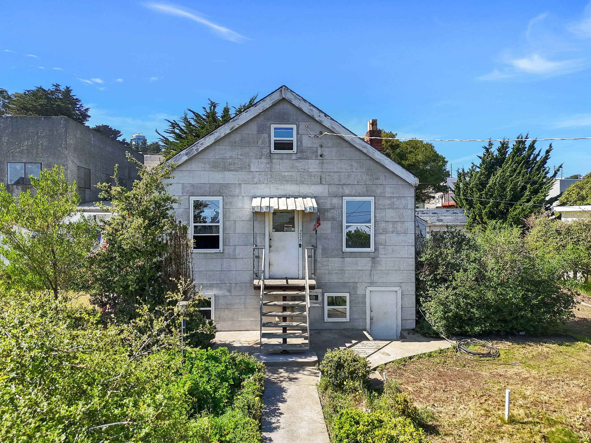 front view of a two-story house with a gable roof, gray siding, and multiple windows. There are stairs leading to a front door with a small awning, and surrounding greenery including bushes and trees under a blue sky.