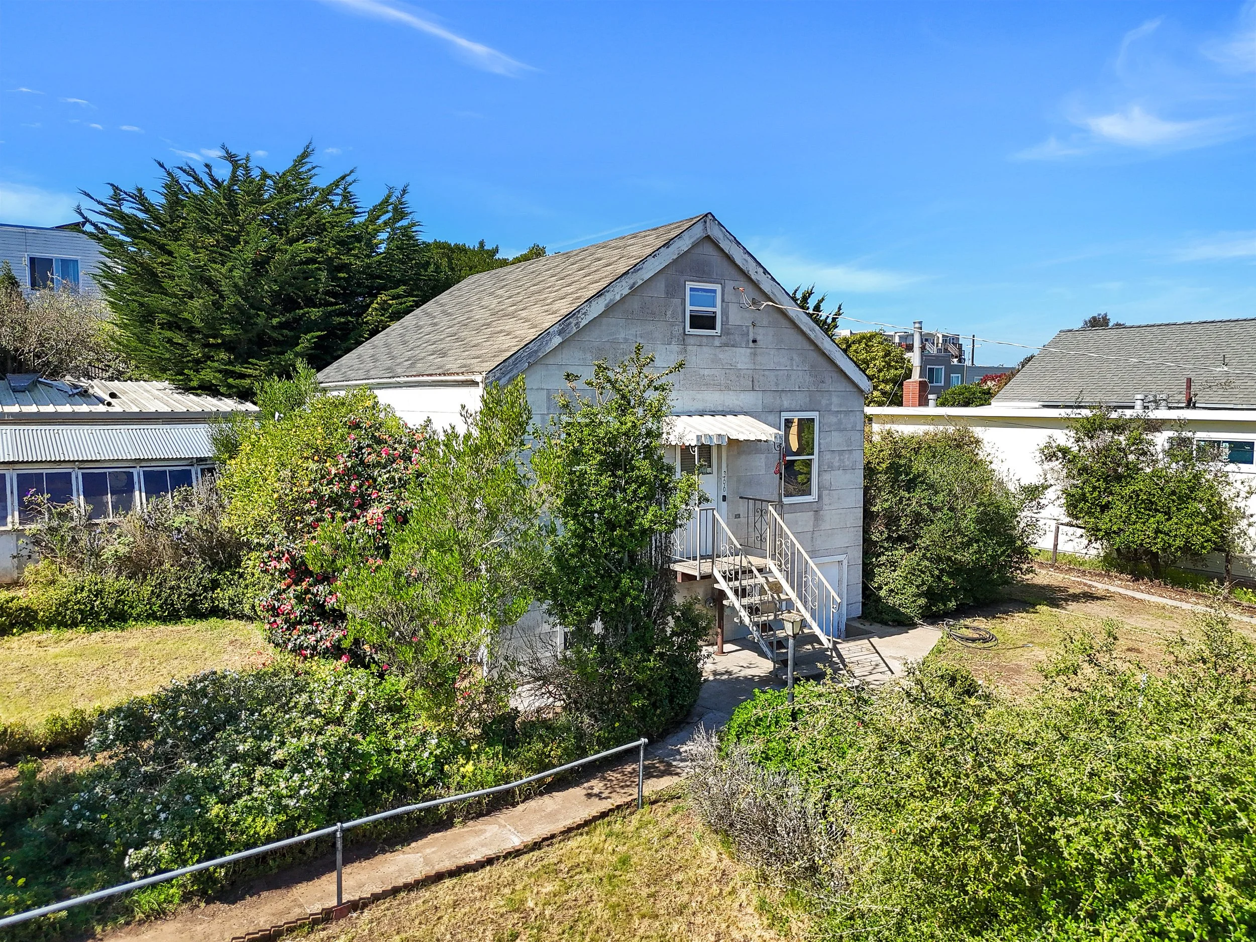A small, gray two-story house with a staircase leading to a front door, surrounded by green bushes and trees, under a bright blue sky.