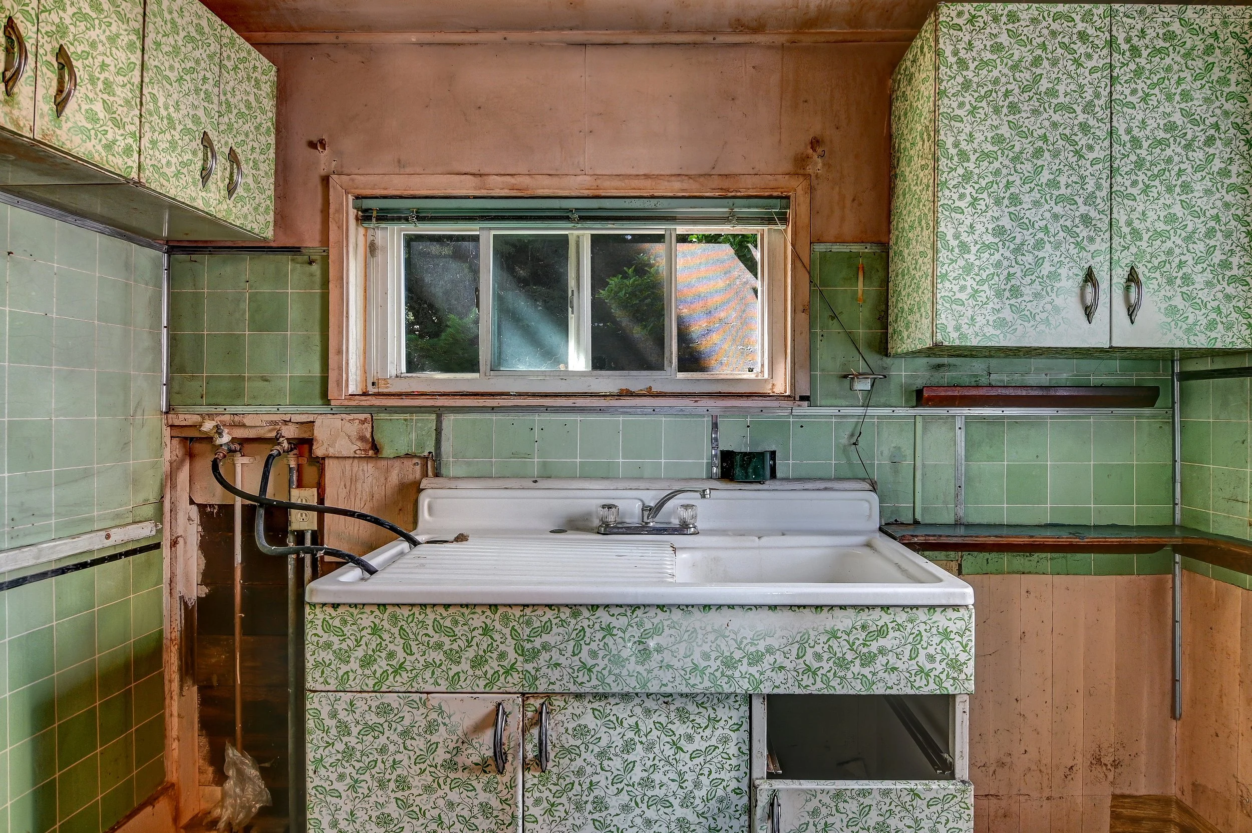 Old kitchen with green patterned cabinets, green tiled walls, and a white sink, showing signs of wear and disrepair.