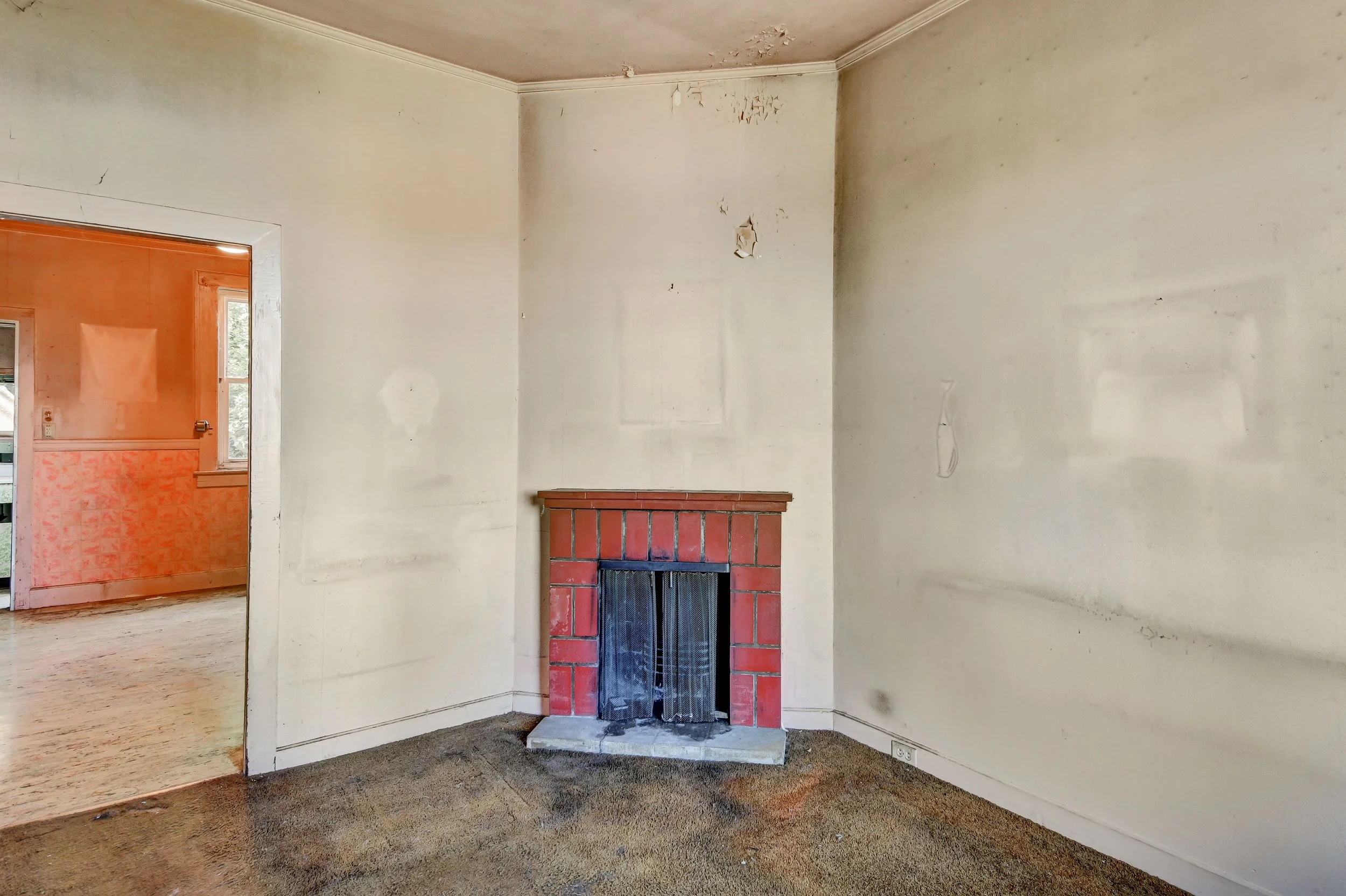 Empty living room with beige walls, a small brick fireplace, and a stained carpet. Part of an adjacent room with orange walls is visible through an open doorway.