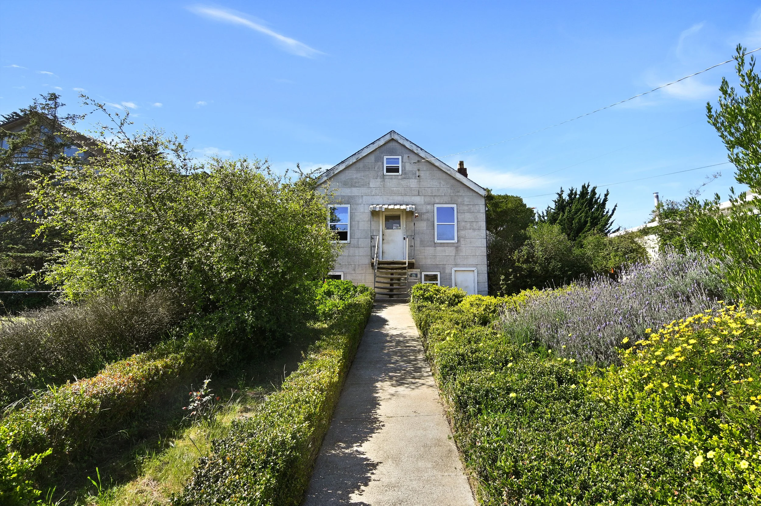A house with a concrete exterior, front steps, overgrown garden, and a clear blue sky.