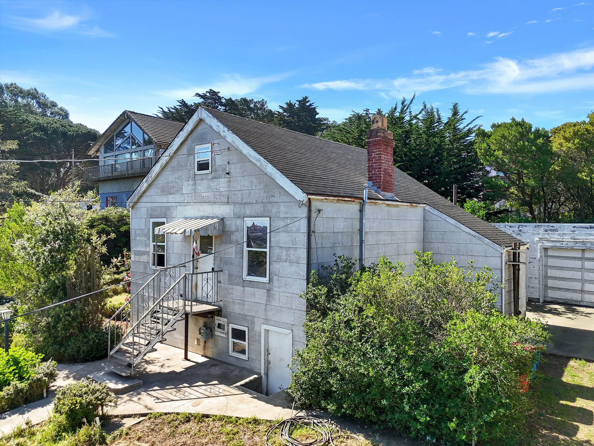 A two-story house with a metal staircase and awning, surrounded by trees and greenery, with a cloudy sky overhead.