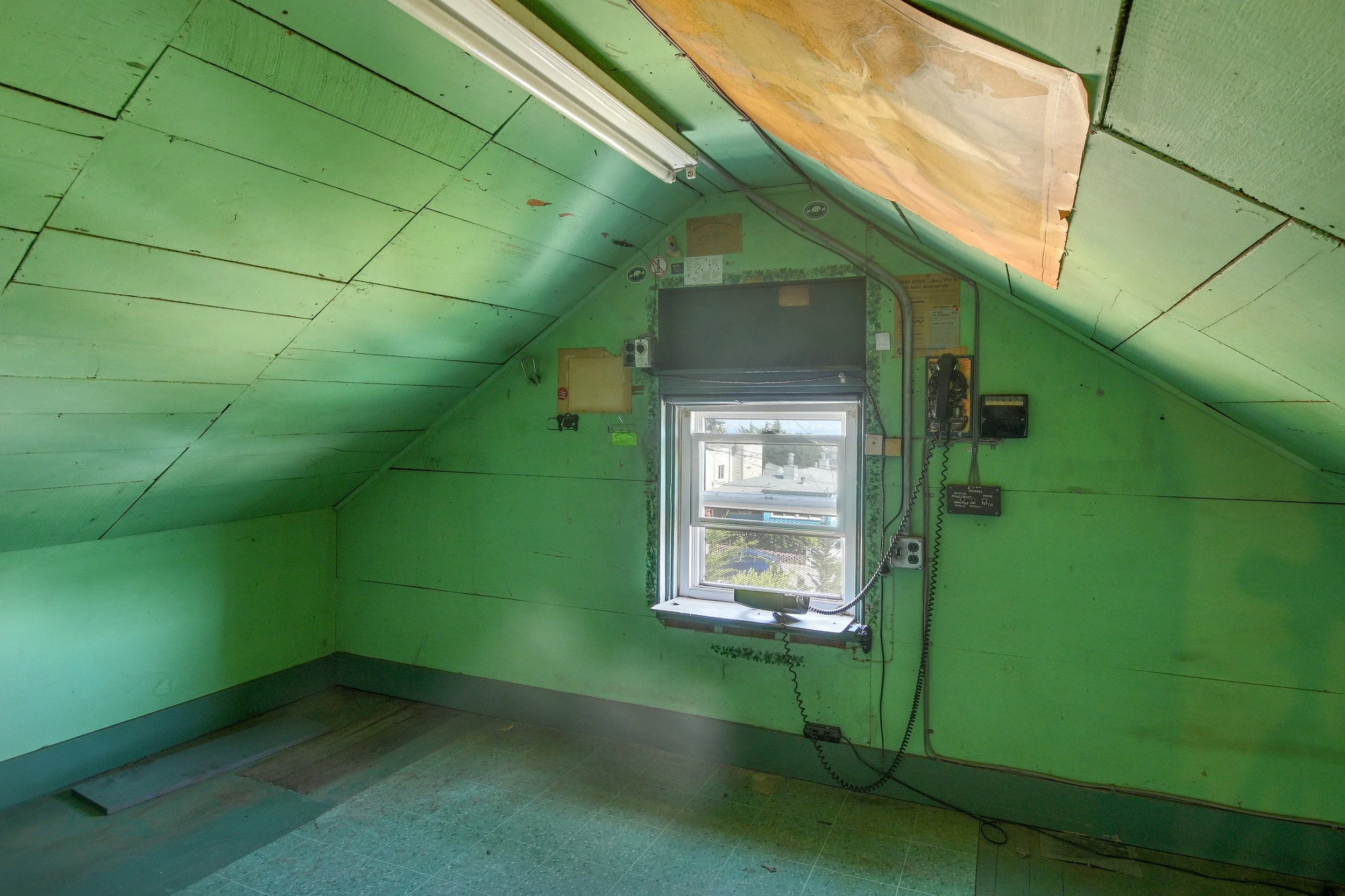An empty room with green painted walls, a small window showing rooftops outside, and various electronic devices and wires attached to the walls. The ceiling is sloped and partially covered with a wooden board.