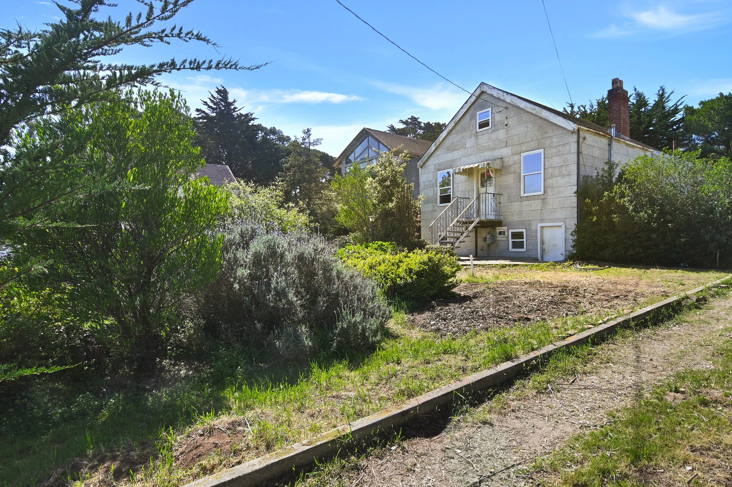 A two-story house with an unfinished exterior, a small front porch with stairs, surrounded by trees and bushes, on a sunny day with a blue sky.