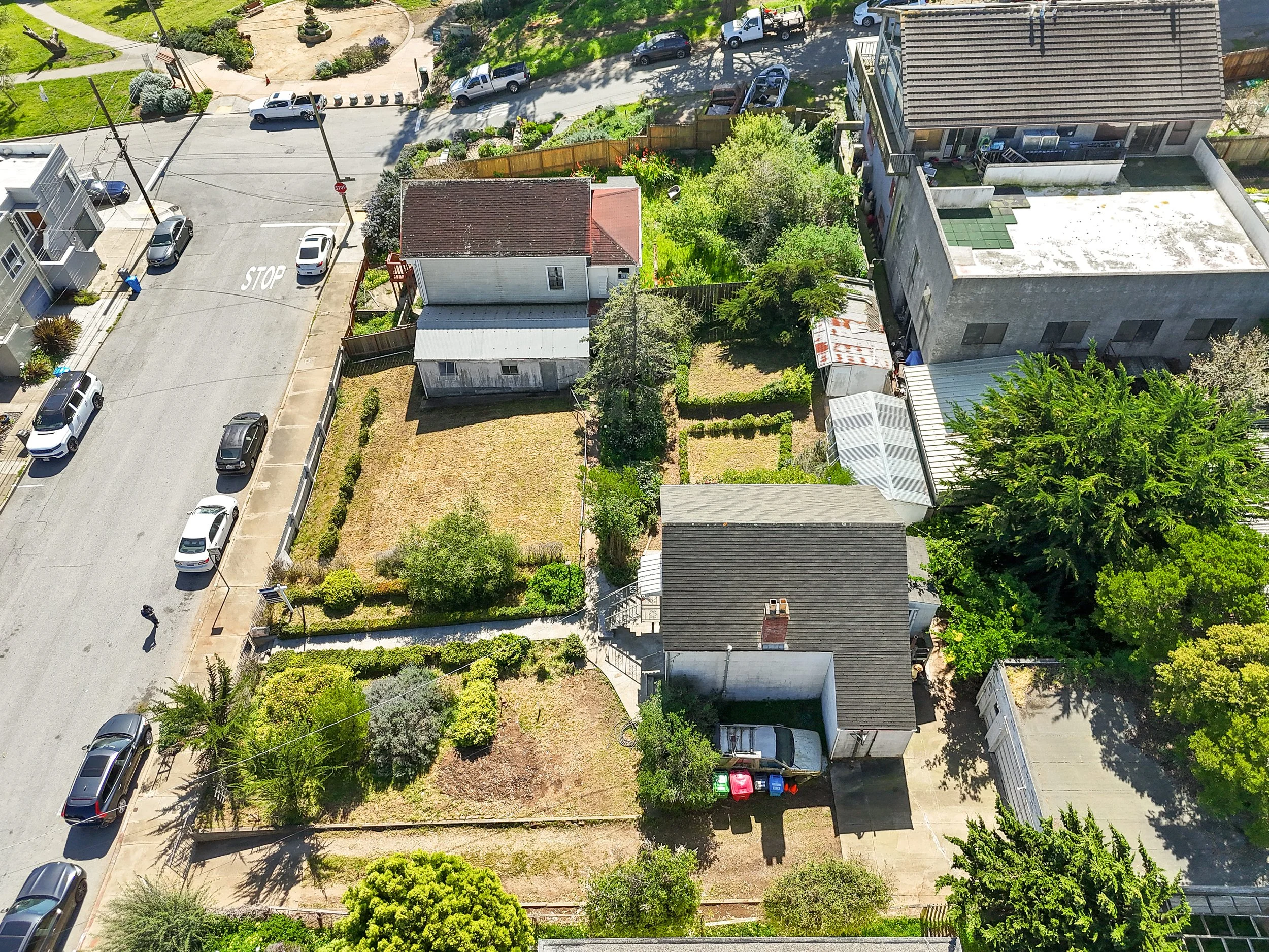 Aerial view of residential neighborhood showing multiple houses, a street with parked cars and a person walking, gardens, trees, and fences.