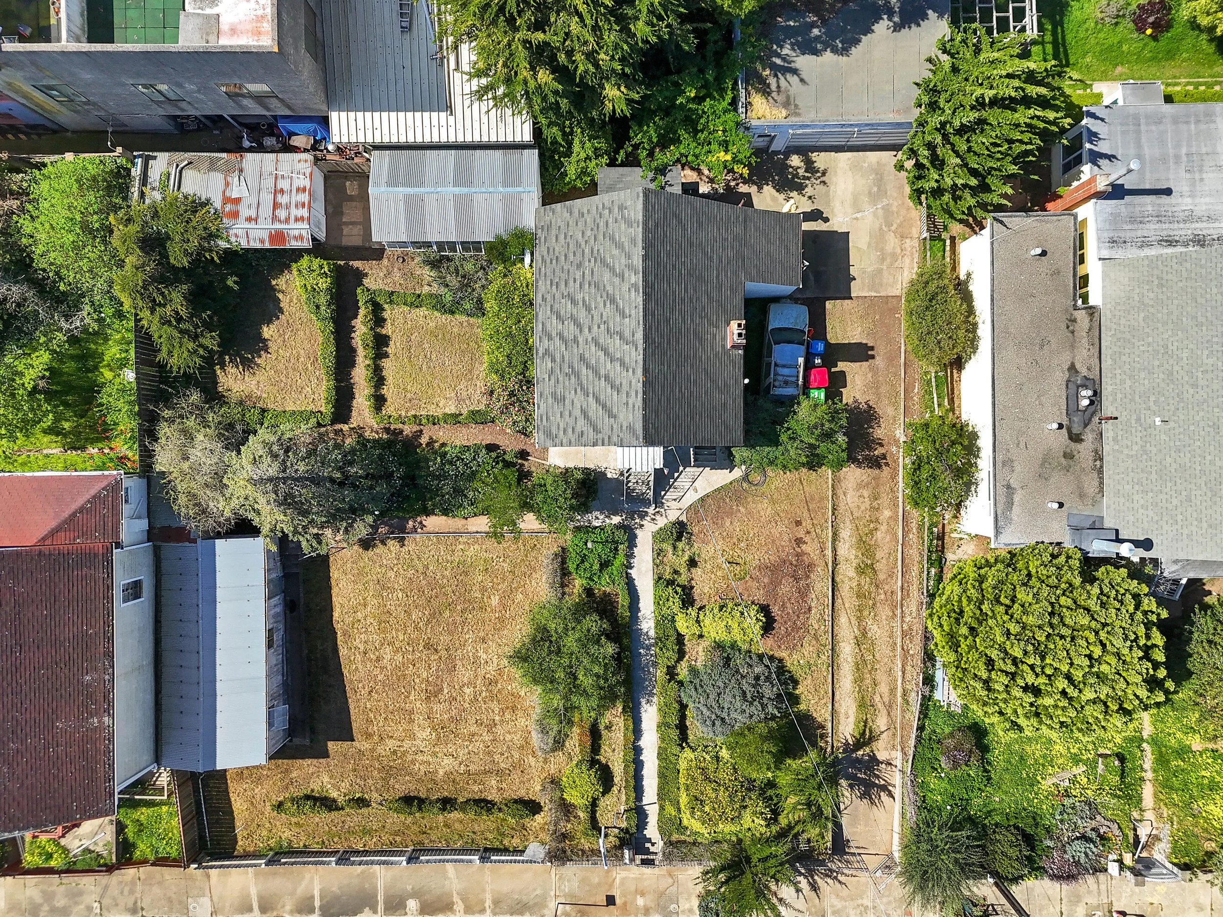 An aerial view of a residential backyard with trees, a small house, and parked cars, surrounded by neighboring houses and yard spaces.