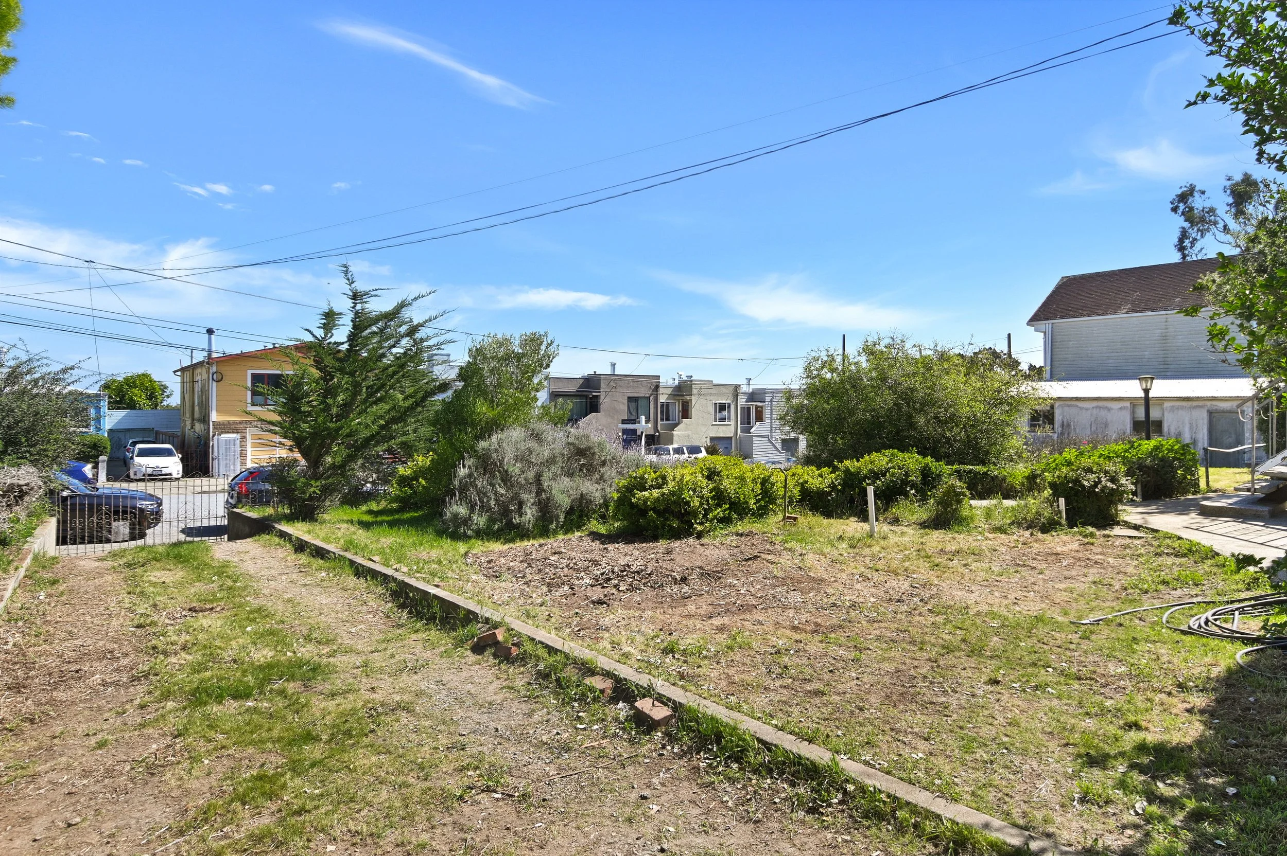 A sunny backyard with a dirt path, a small garden bed, and bushes. In the background, there are several houses and trees, with a clear blue sky overhead.