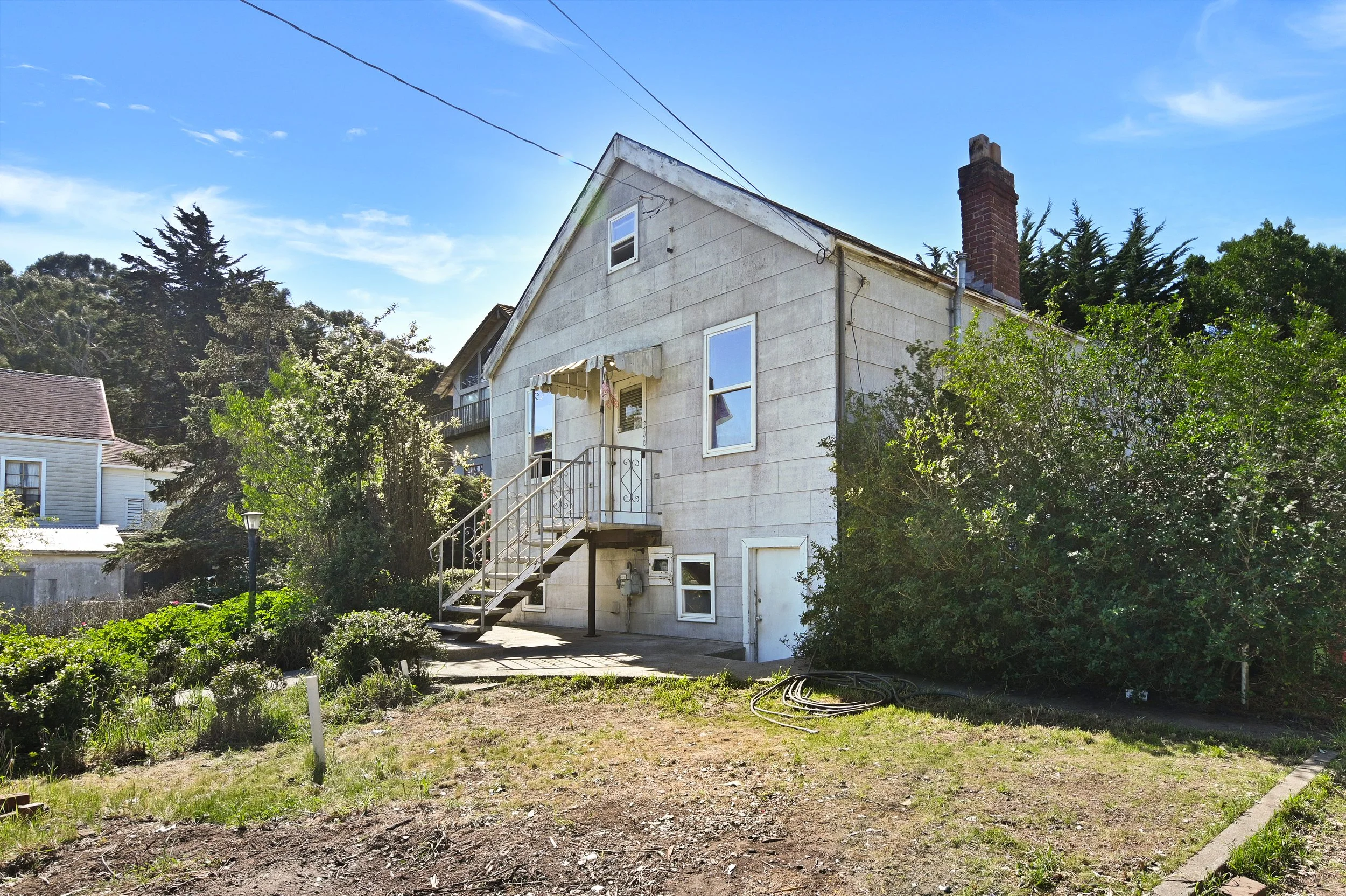 Back of a two-story house with white siding, a brick chimney, an outdoor staircase, and a small backyard with bushes and trees under a blue sky.