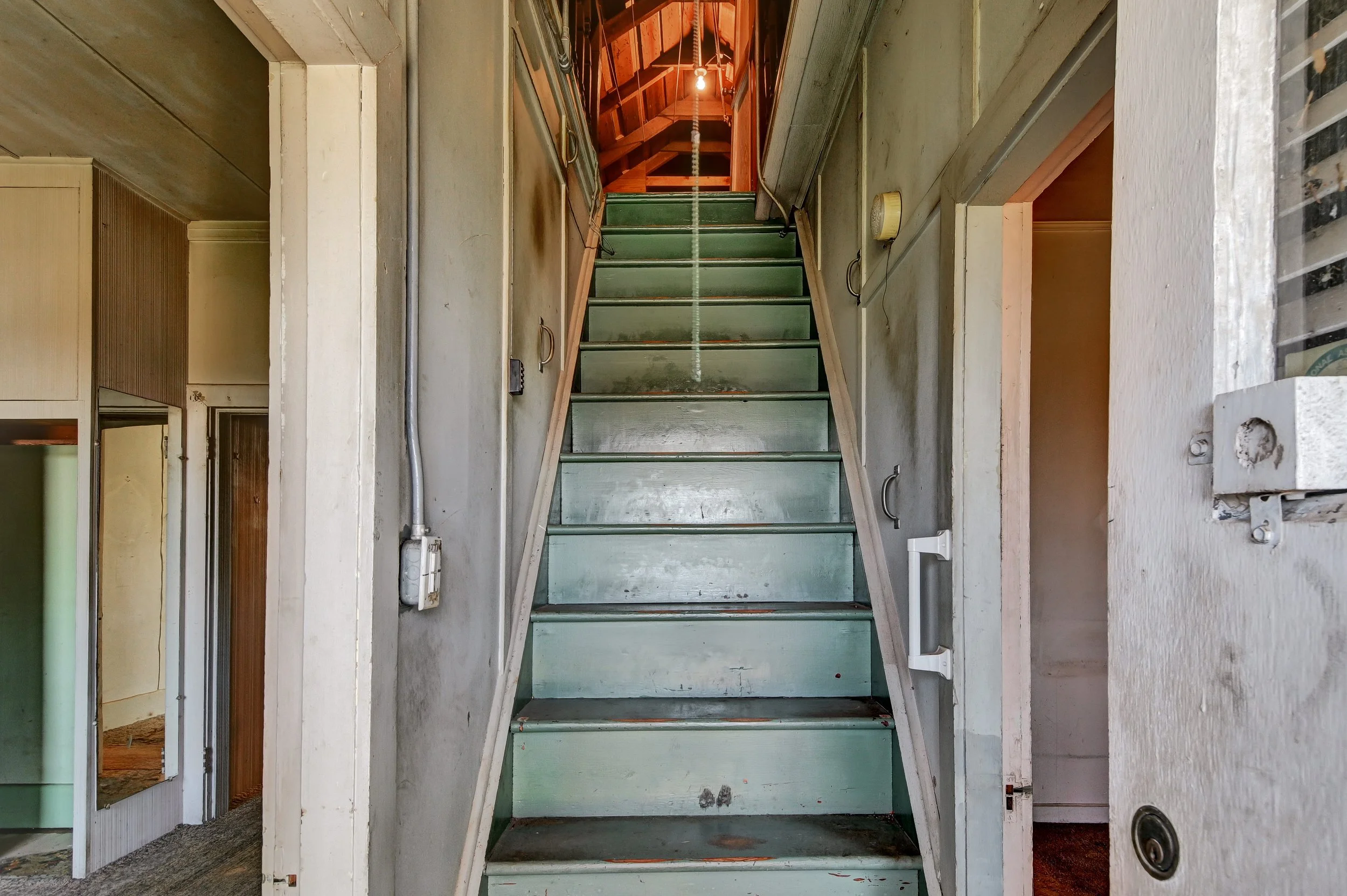 Interior of an old, weathered house with a staircase leading to an upper floor. The stairs are painted light green and look worn. To the left, there is a doorway with a mirror, and to the right, a wall with visible signs of age. The ceiling at the to