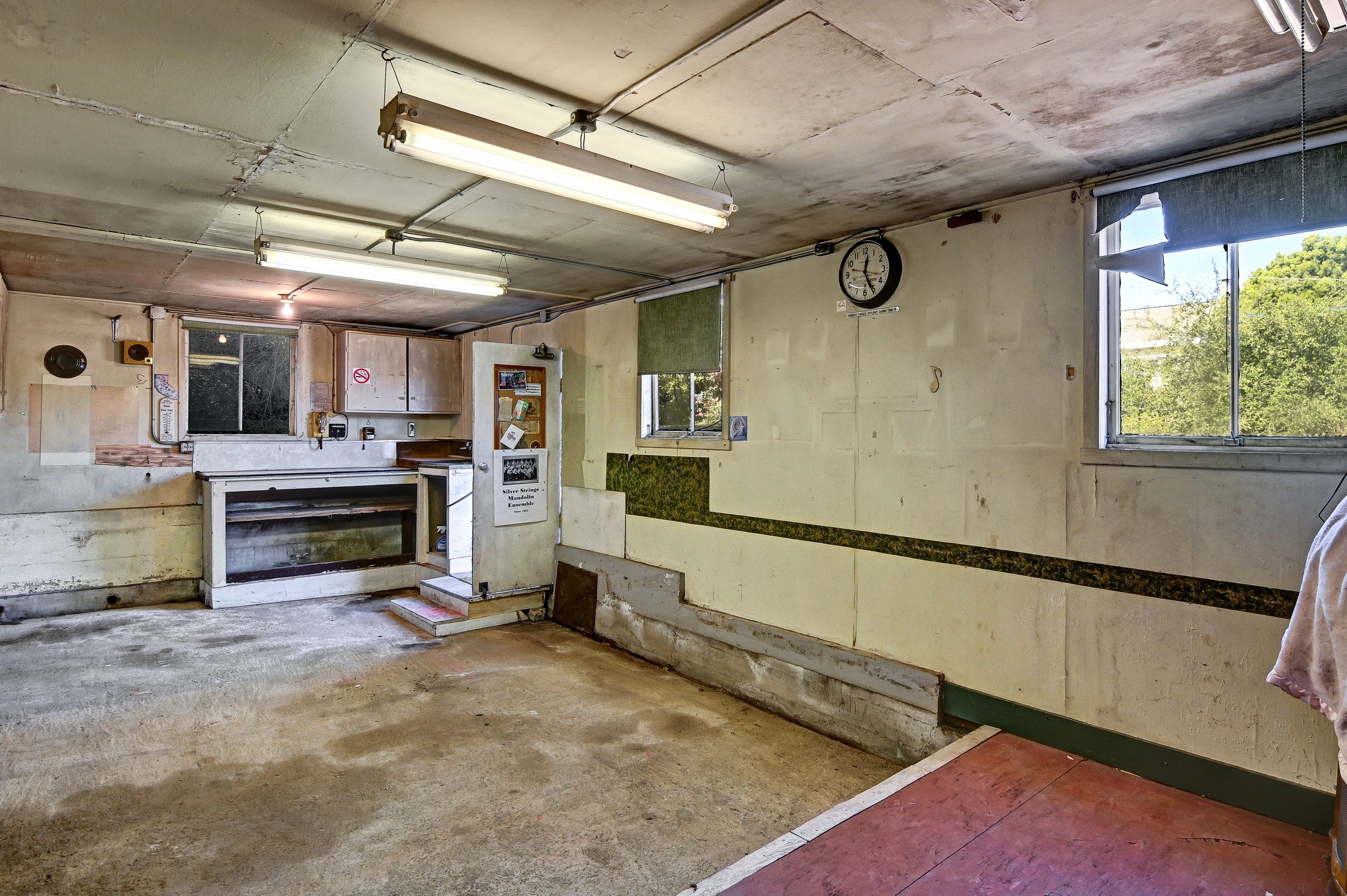 Empty, worn-out garage with peeling walls and stained concrete floor. Fluorescent lights on the ceiling, small windows with blinds, a clock on the wall, and a cluttered workbench area.
