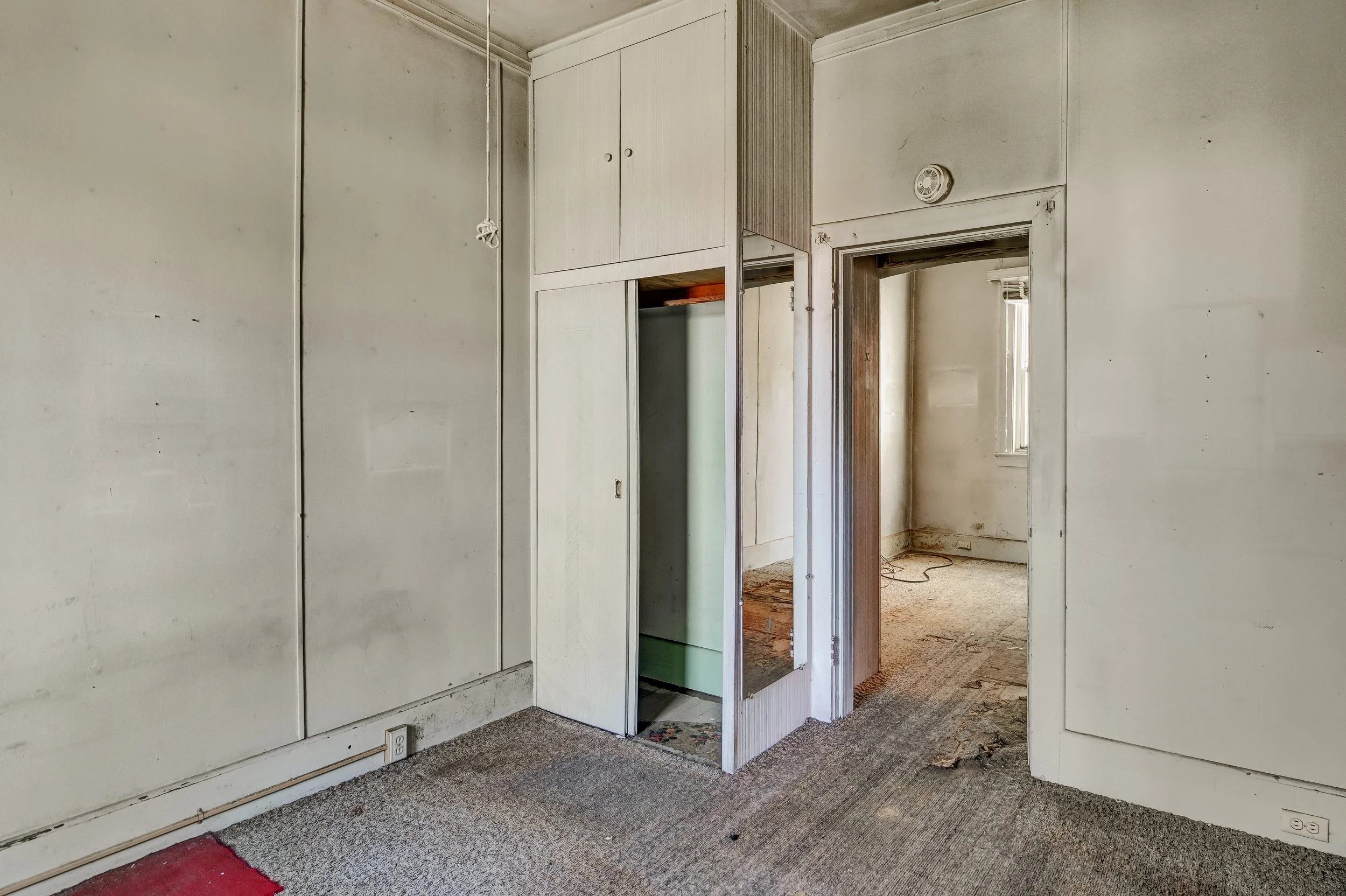 Room with beige walls and old carpet, small closet with doors, and an open doorway leading to a second room with a window and damaged flooring.