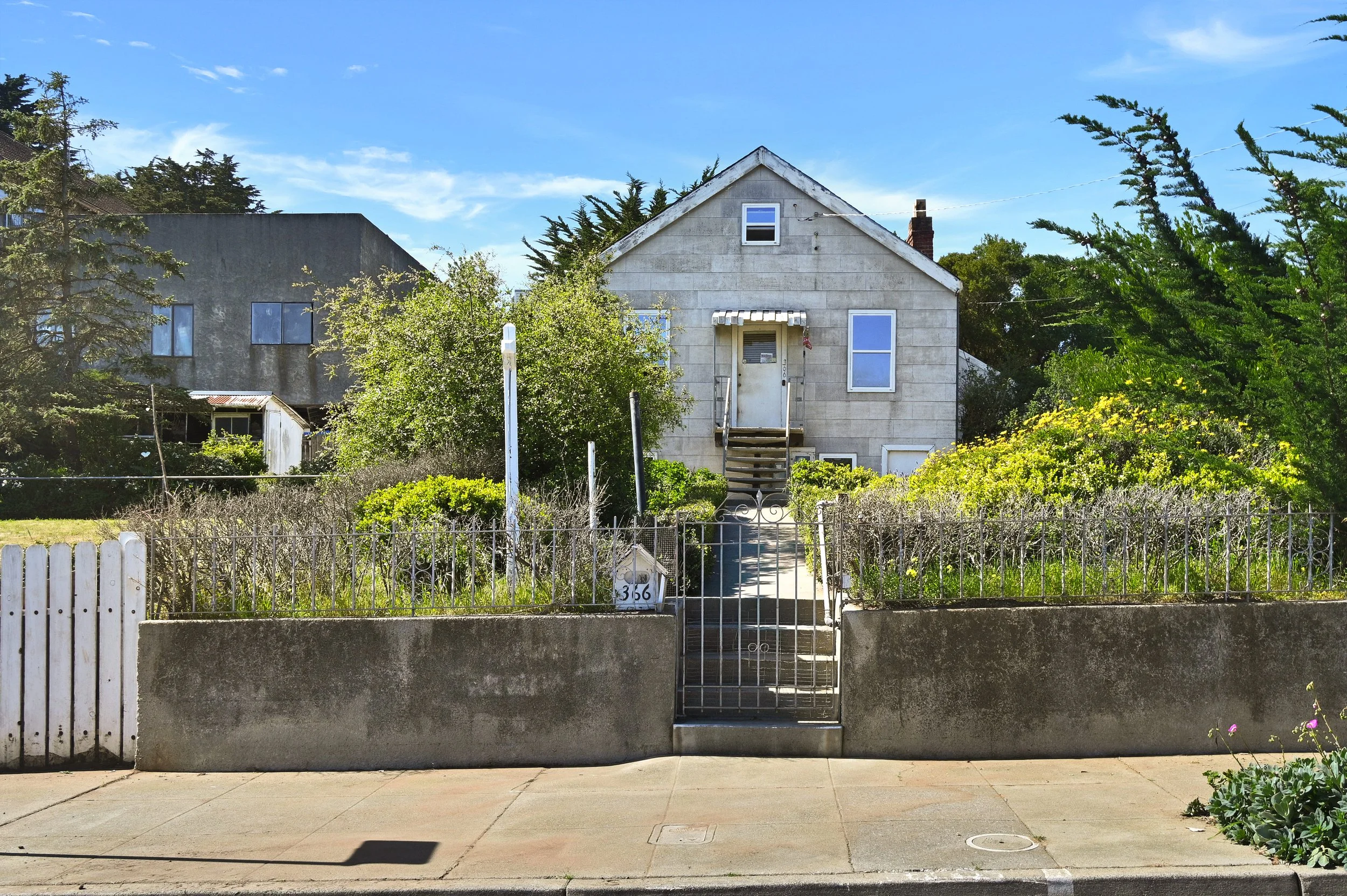 A two-story house with gray exterior, multiple windows, front steps, and a small covered porch, surrounded by greenery and a garden with a metal fence and sidewalk in front.