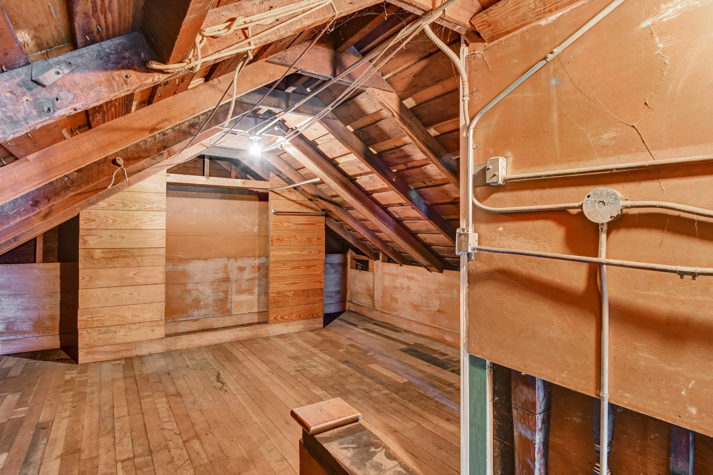 Interior view of an attic with unfinished wood walls, ceiling, and floor. Visible electrical wiring and a central light fixture.