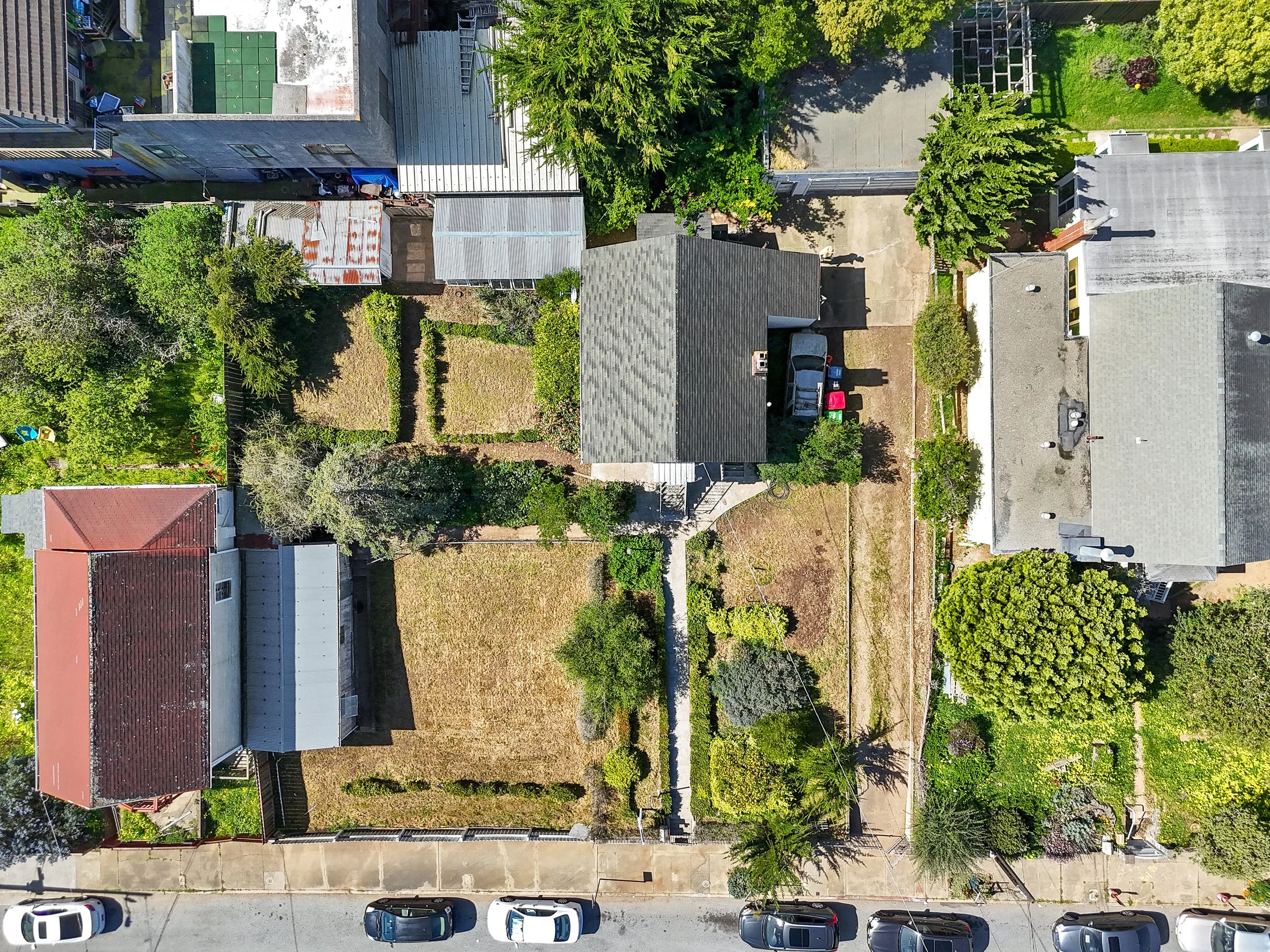An aerial view of residential houses, trees, gardens, driveways, and parked cars in a neighborhood.