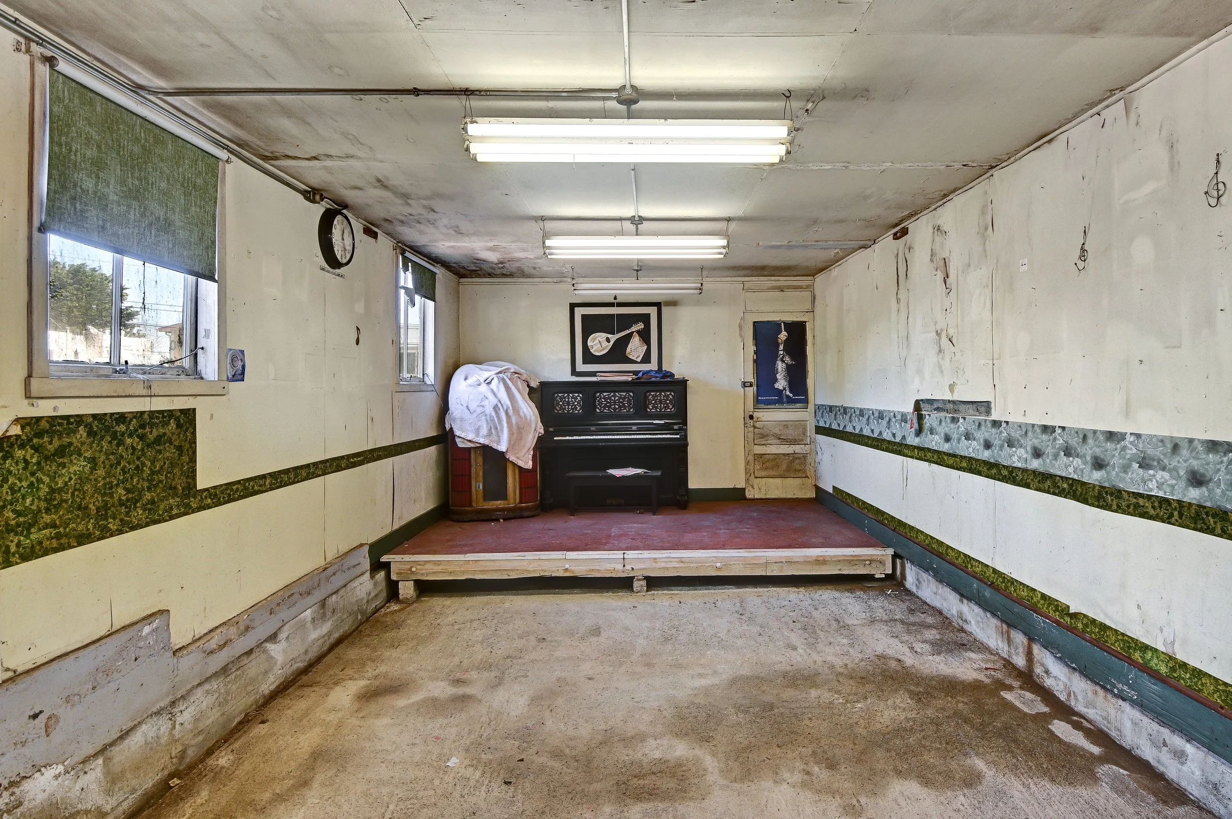 Empty, worn-down room with peeling walls, two small windows with green blinds, fluorescent ceiling lights, and a small raised stage with a black upright piano topped with a wrapped object. A framed picture of musical instruments hangs on the back wal