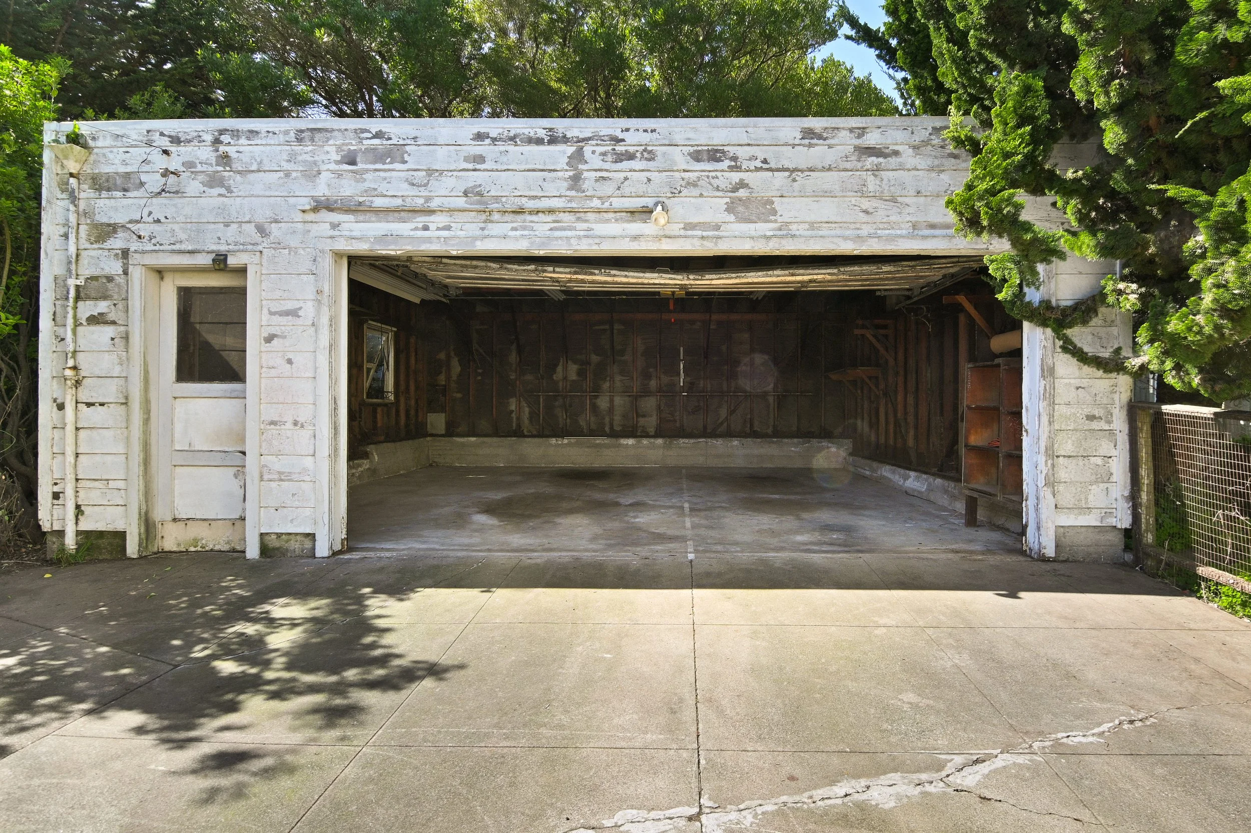 Empty, weathered garage with peeling white paint, open door, concrete floor, and wooden walls, surrounded by trees and greenery.