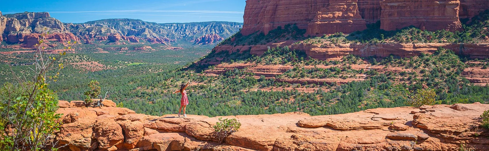 A person in a red dress standing on a rocky ledge in a desert landscape with red rock formations and green vegetation.