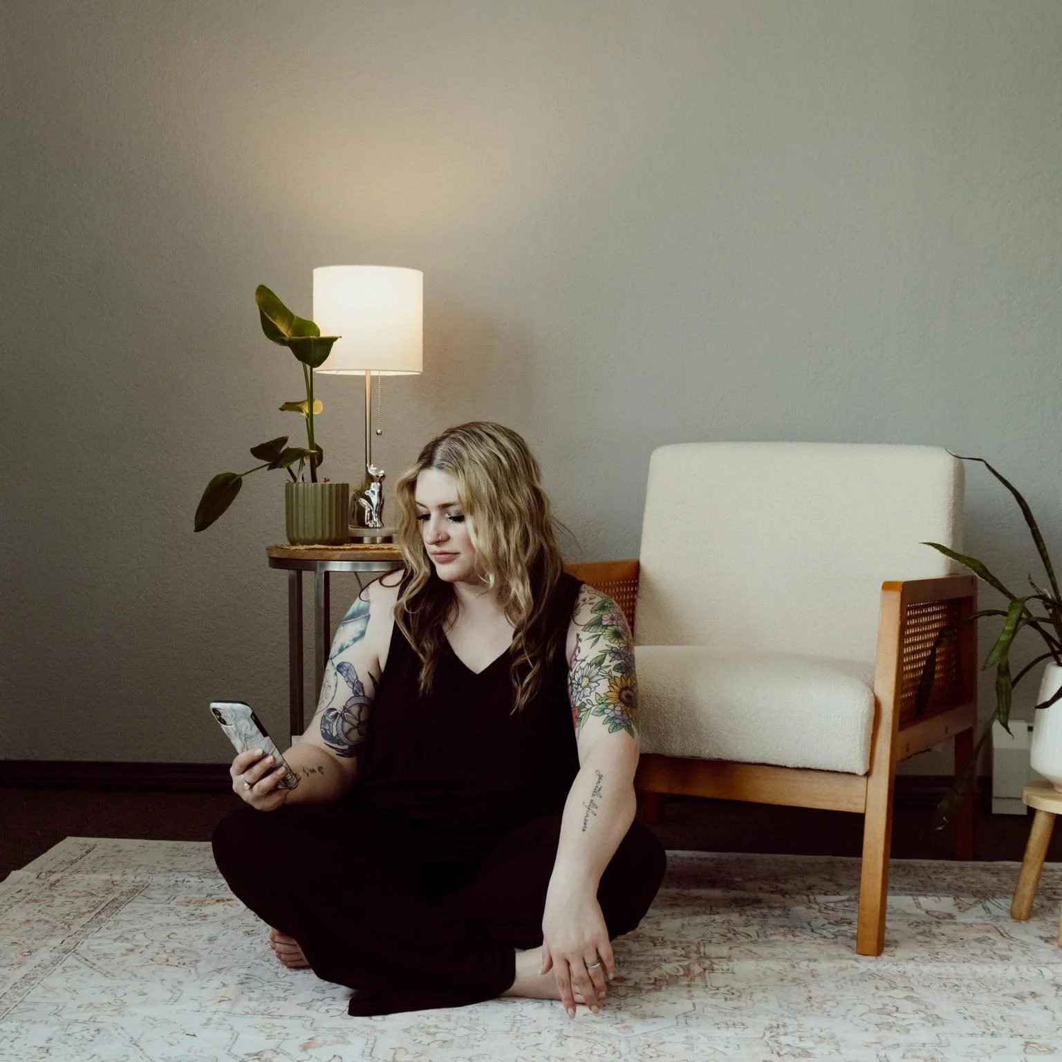 A woman with tattoos sitting cross-legged on a rug in a room, looking at her phone. There's a cream-colored armchair, a side table with a lamp, and a potted plant in the background.