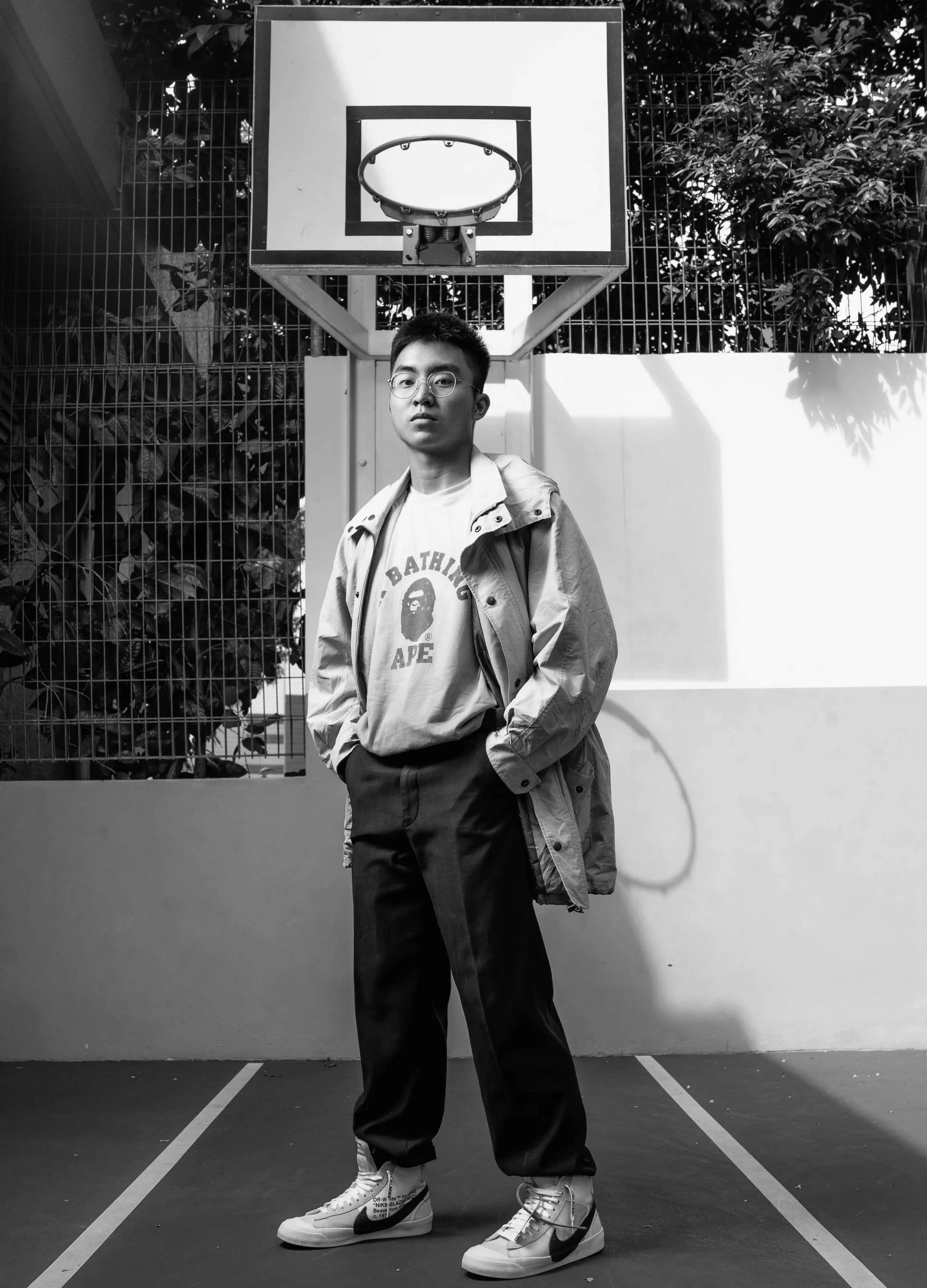 A young man standing on a basketball court with a basketball hoop behind him. He is wearing glasses, a graphic T-shirt, a jacket, and sneakers, with one hand in his pocket.