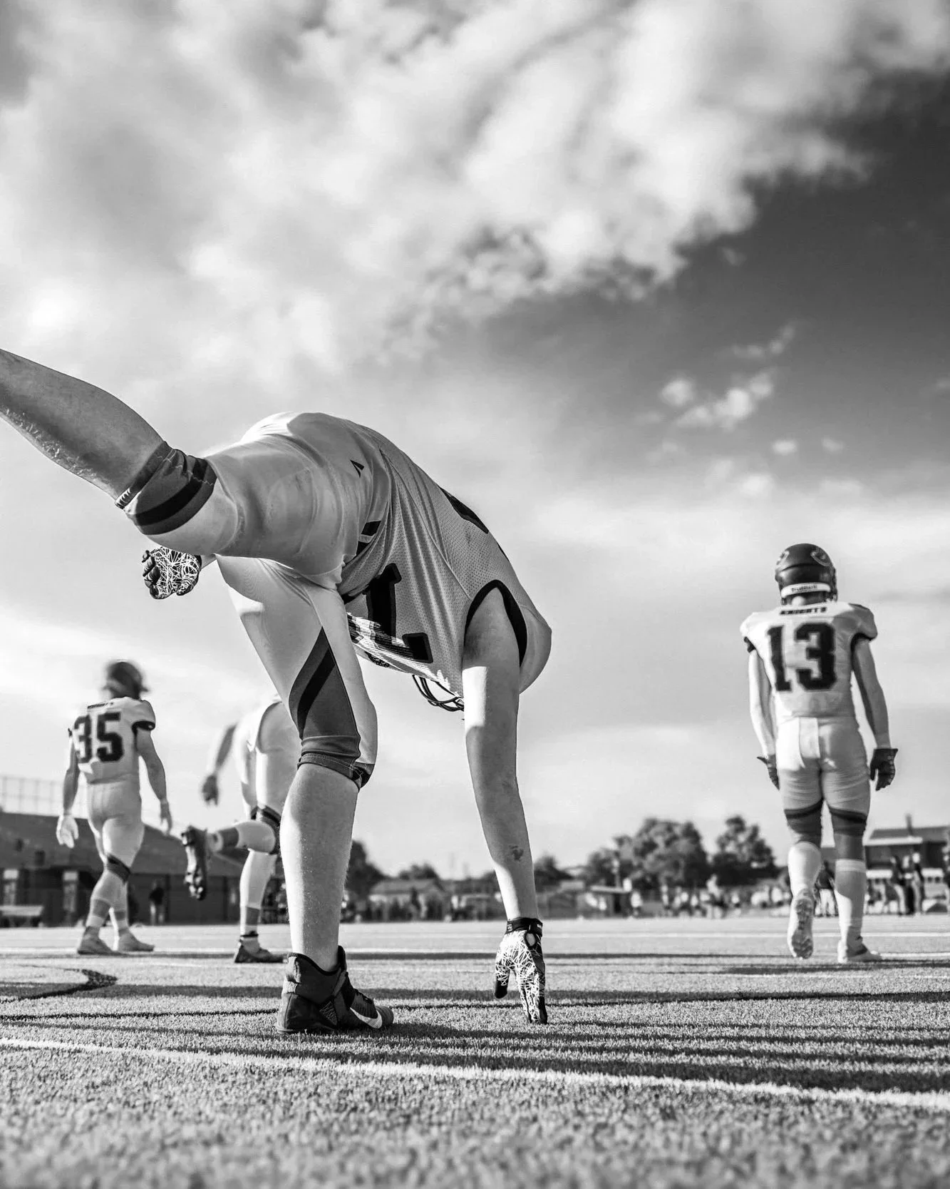 Football players on the field during a game, with one player in foreground reaching for the ground and others walking or standing in the background, black and white photo.