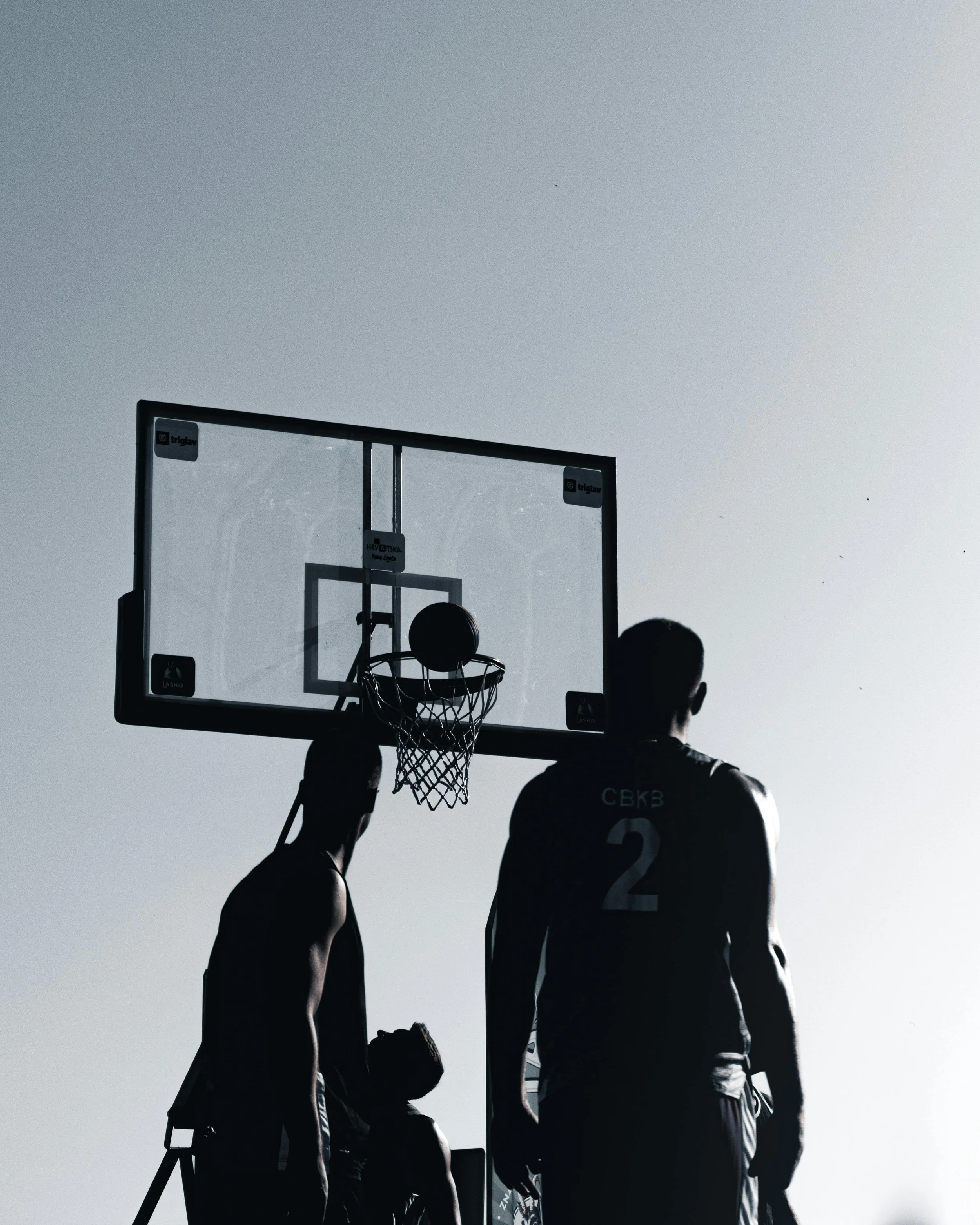 Silhouettes of basketball players standing under a basketball hoop during daytime, with the sky in the background.