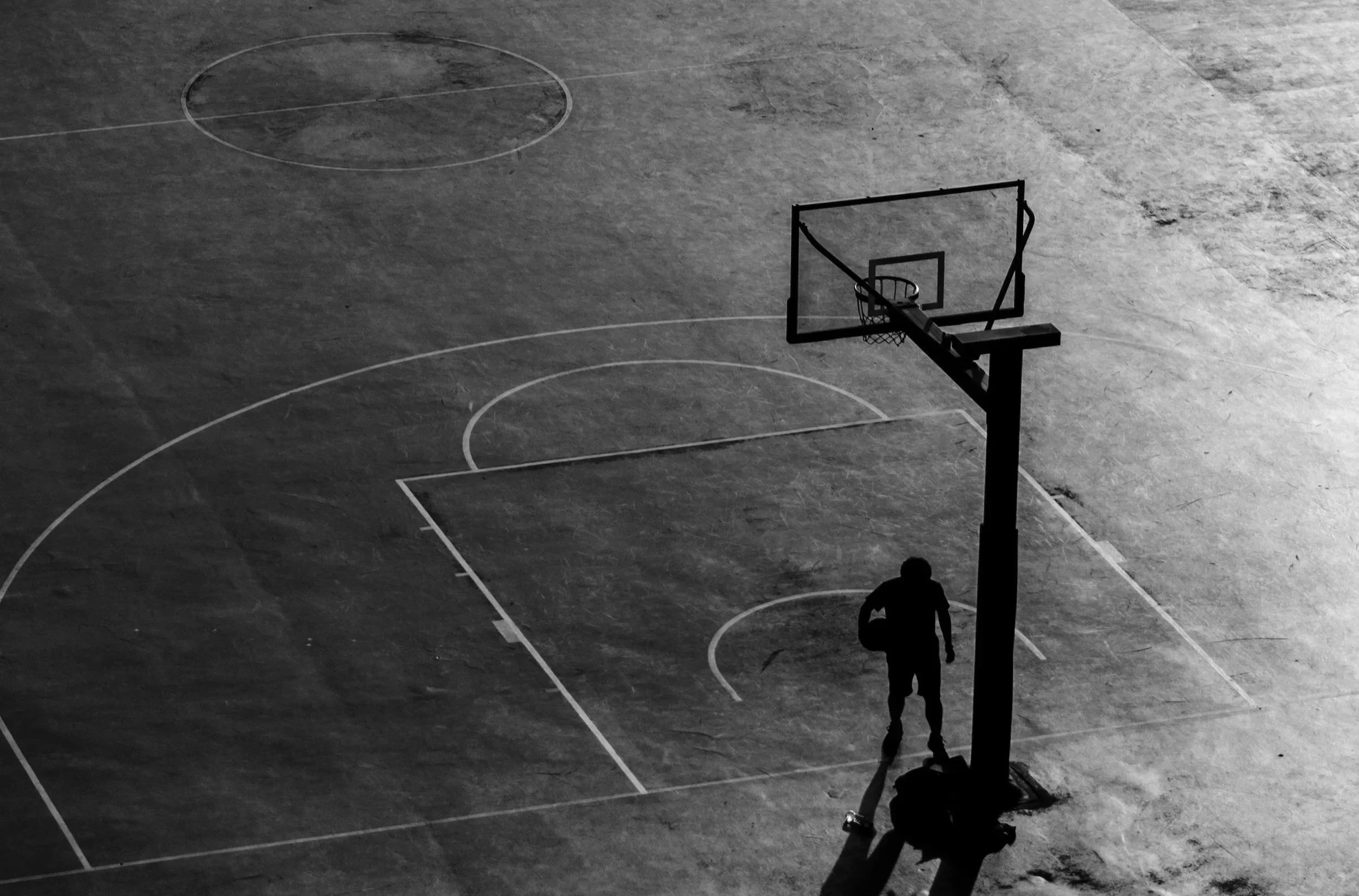 A silhouette of a person standing on an outdoor basketball court at night. The person is holding a basketball and purple sneakers are visible. The court is empty, and the scene is lit by a single light source, creating a long shadow of the person.