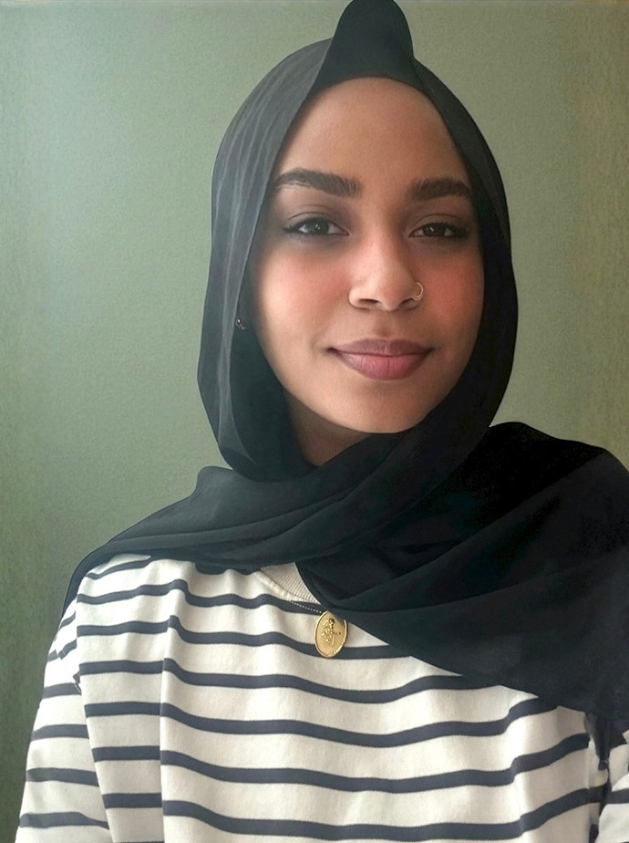 A woman wearing a black headscarf, striped shirt, and a gold pendant, smiling with a green background.