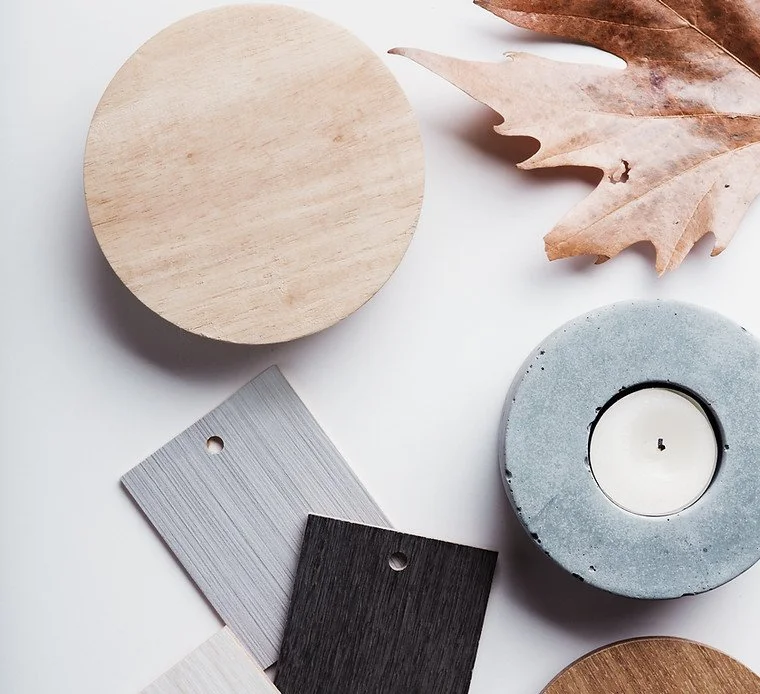 Flat lay of home decor items including a round wooden coaster, a grey and black paint sample, a candle in a concrete holder, and a dried brown leaf.